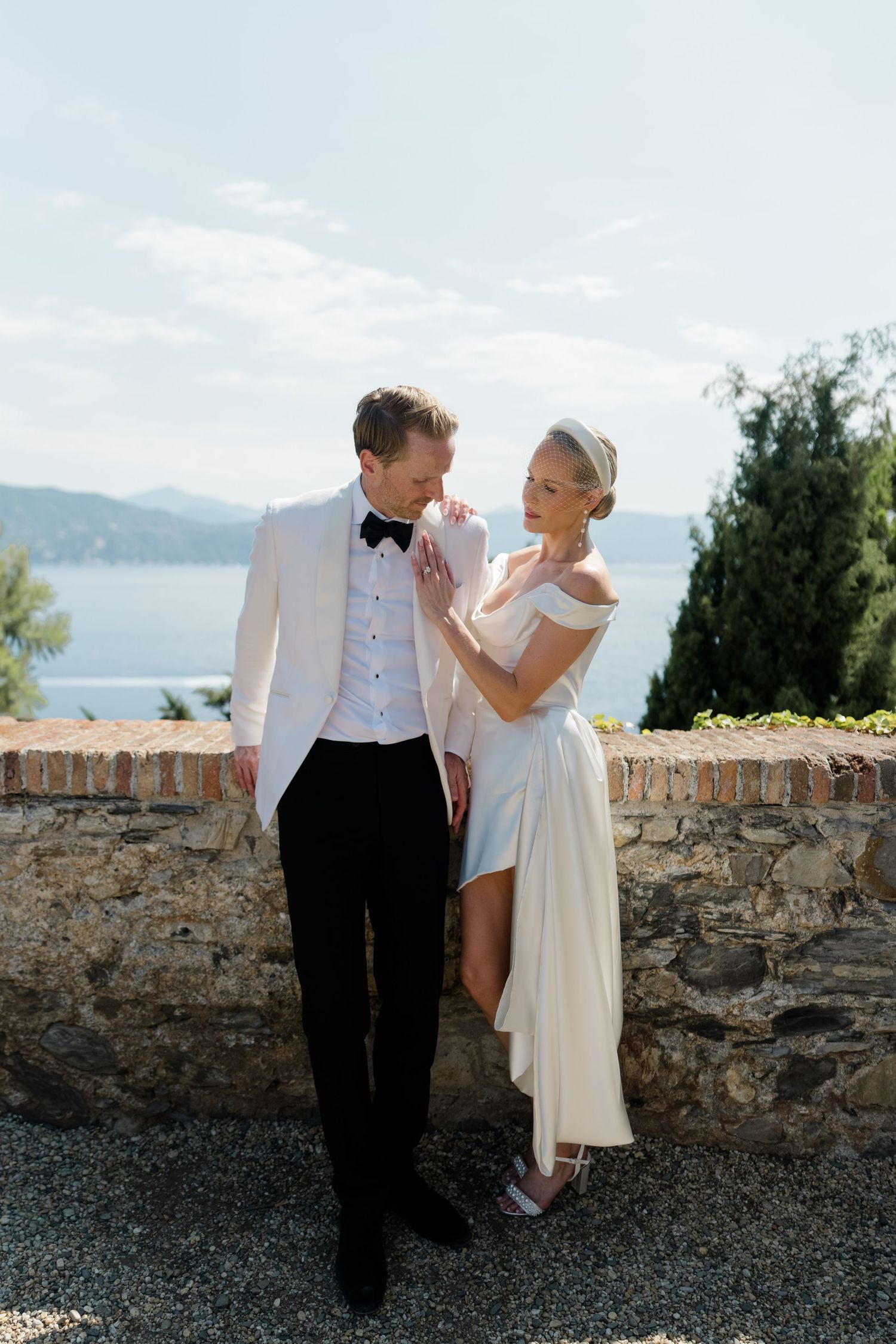Elegant couple in wedding attire share romantic moment by stone wall with scenic lake and mountain backdrop.