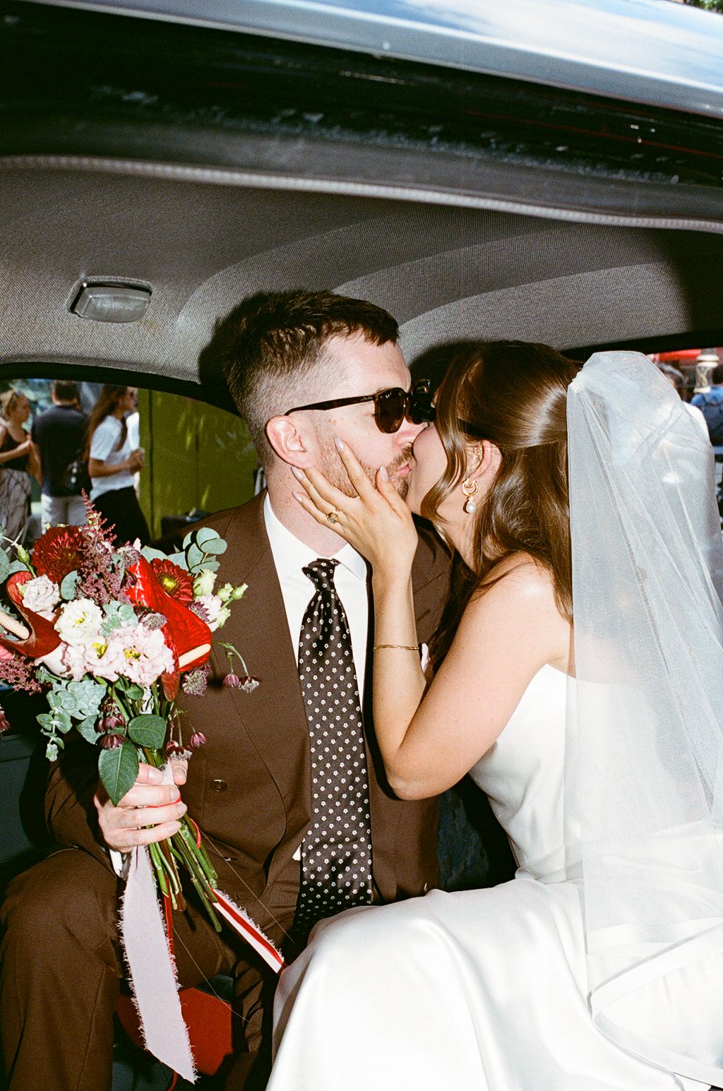Newlyweds kissing in car backseat, groom in brown suit holding red and white bridal bouquet.