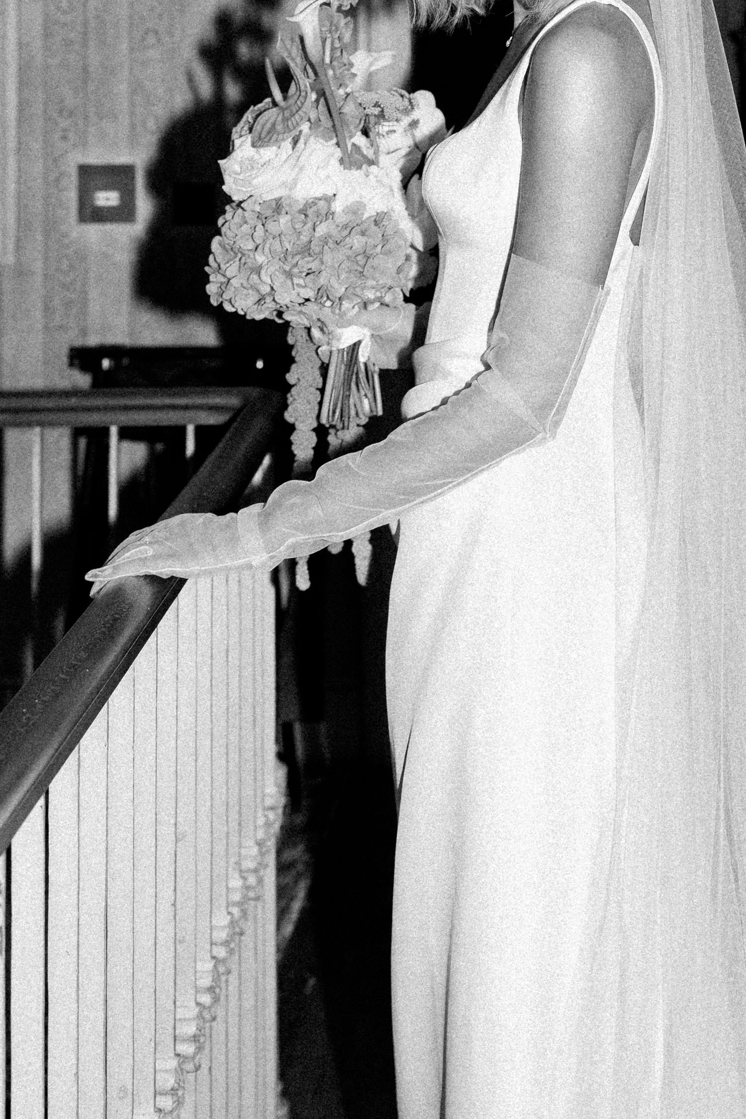 Black and white photo of a bride in a white gown and veil holding a bouquet while standing at a staircase railing.