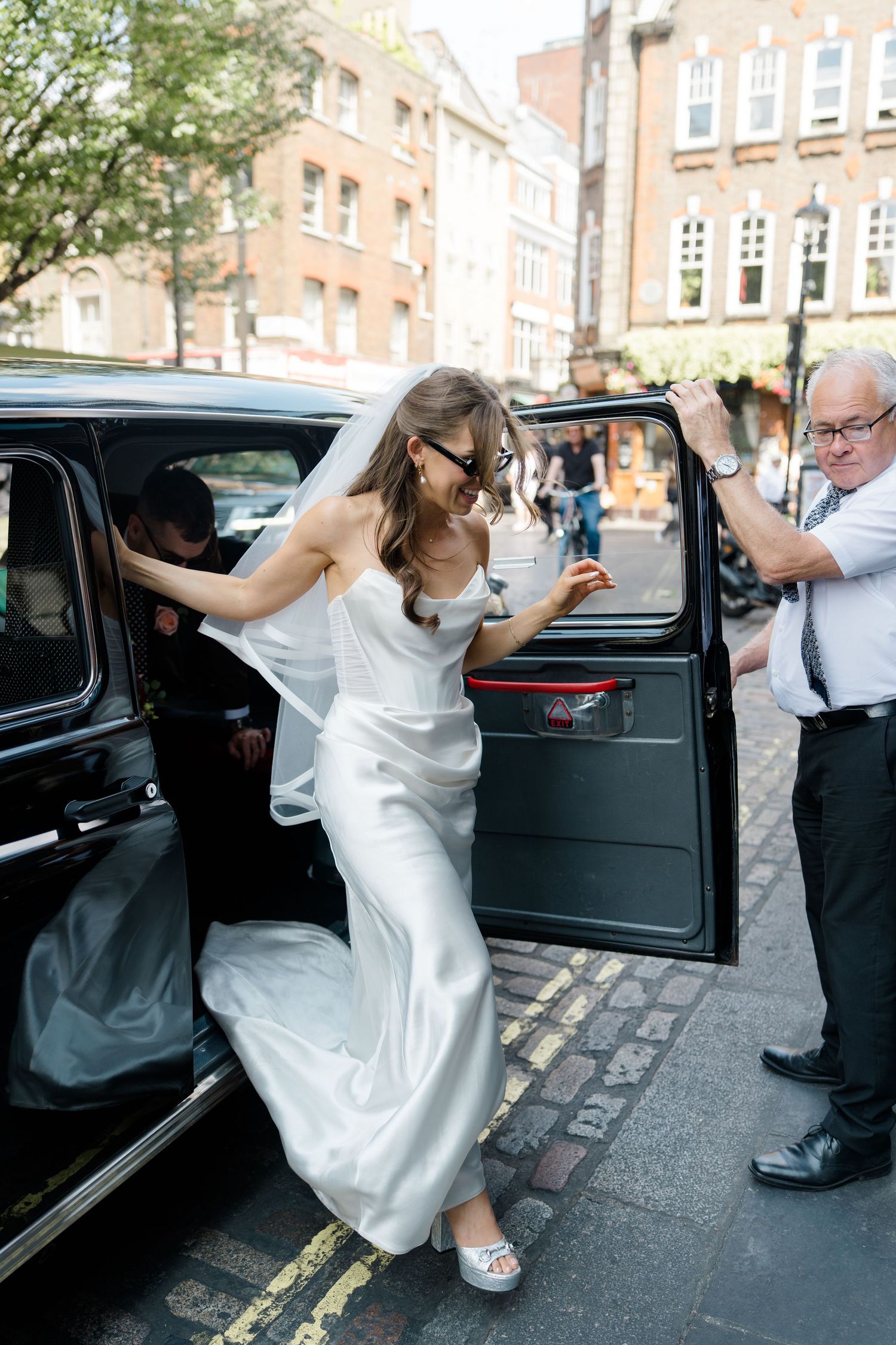 Bride in white satin wedding dress and veil stepping out of a black London taxi cab.