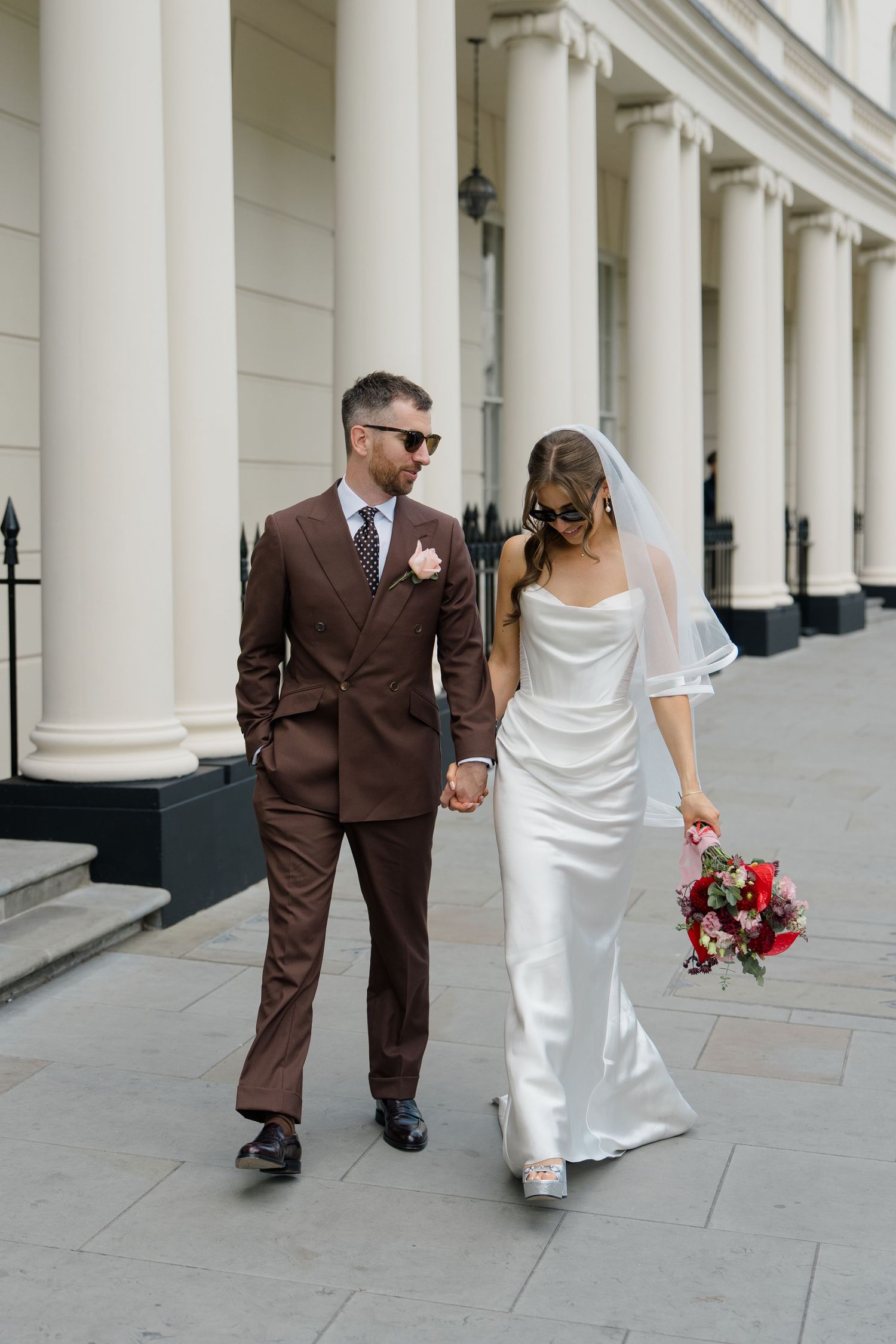 Stylish groom in brown suit and bride in white satin gown walking past white columns holding bouquet.