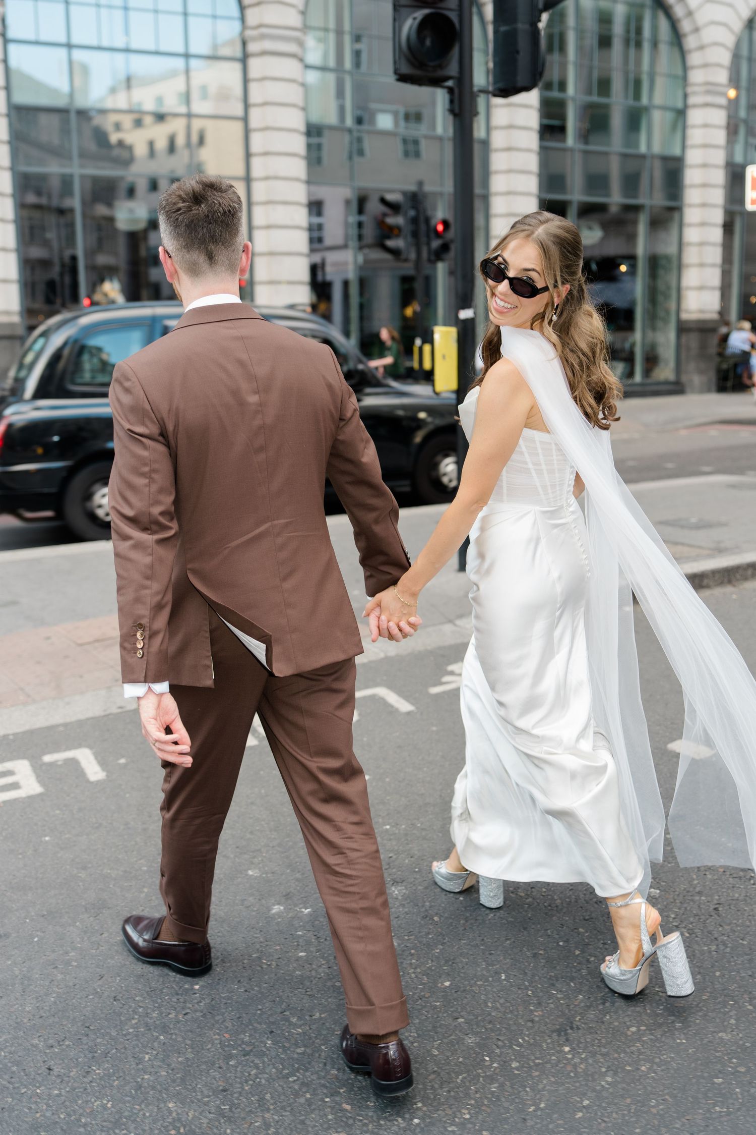 Bride and groom in wedding attire holding hands while crossing a London city street.