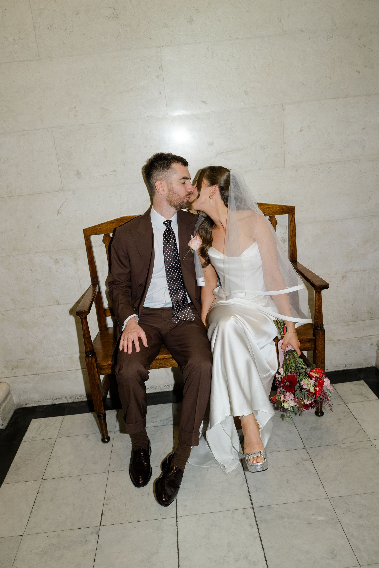 Bride and groom kiss while seated on a wooden bench, bride holding a red and pink bouquet.
