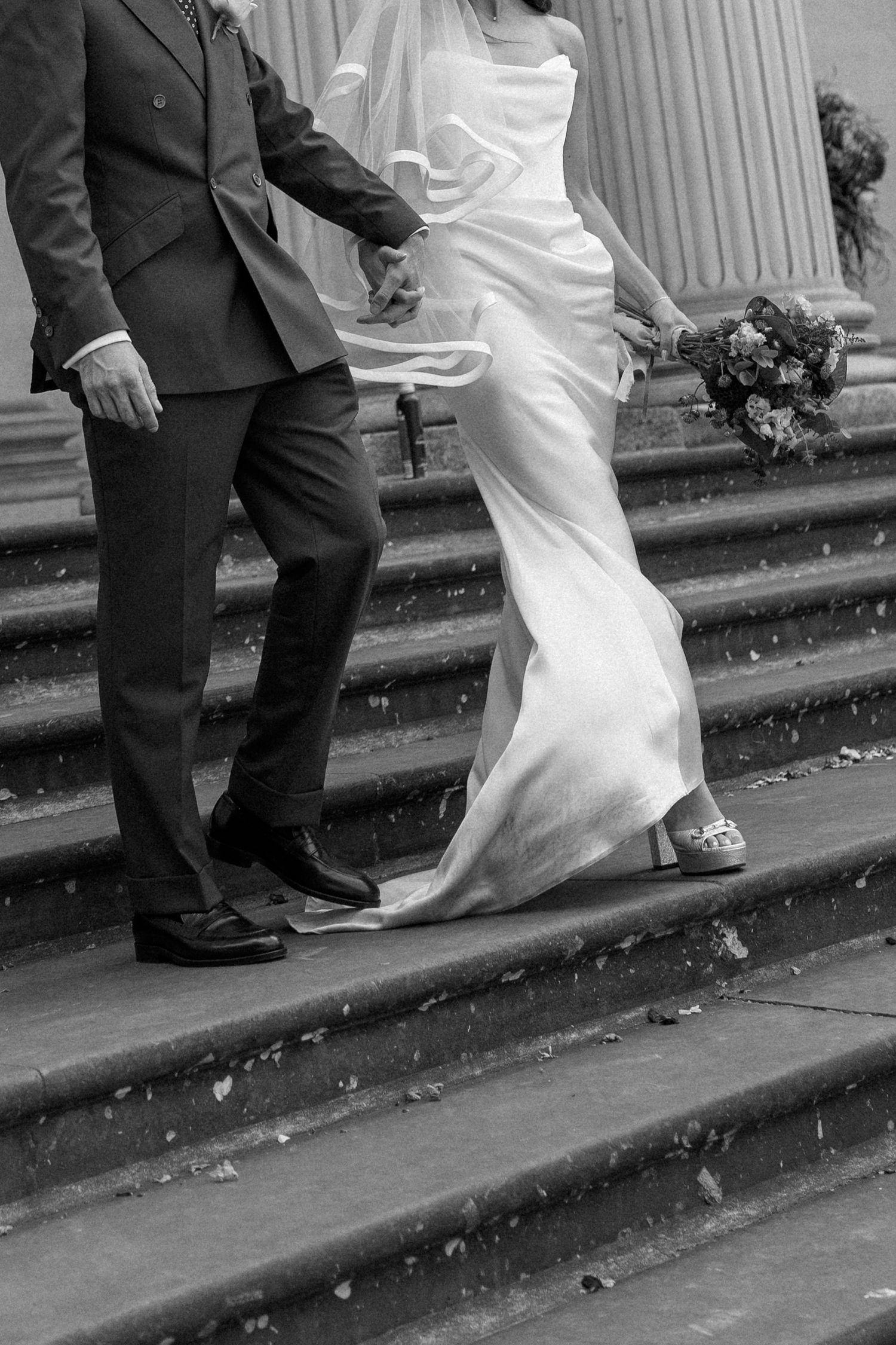 Bride and groom walk down stone steps hand in hand, veil flowing, in elegant black and white photo.