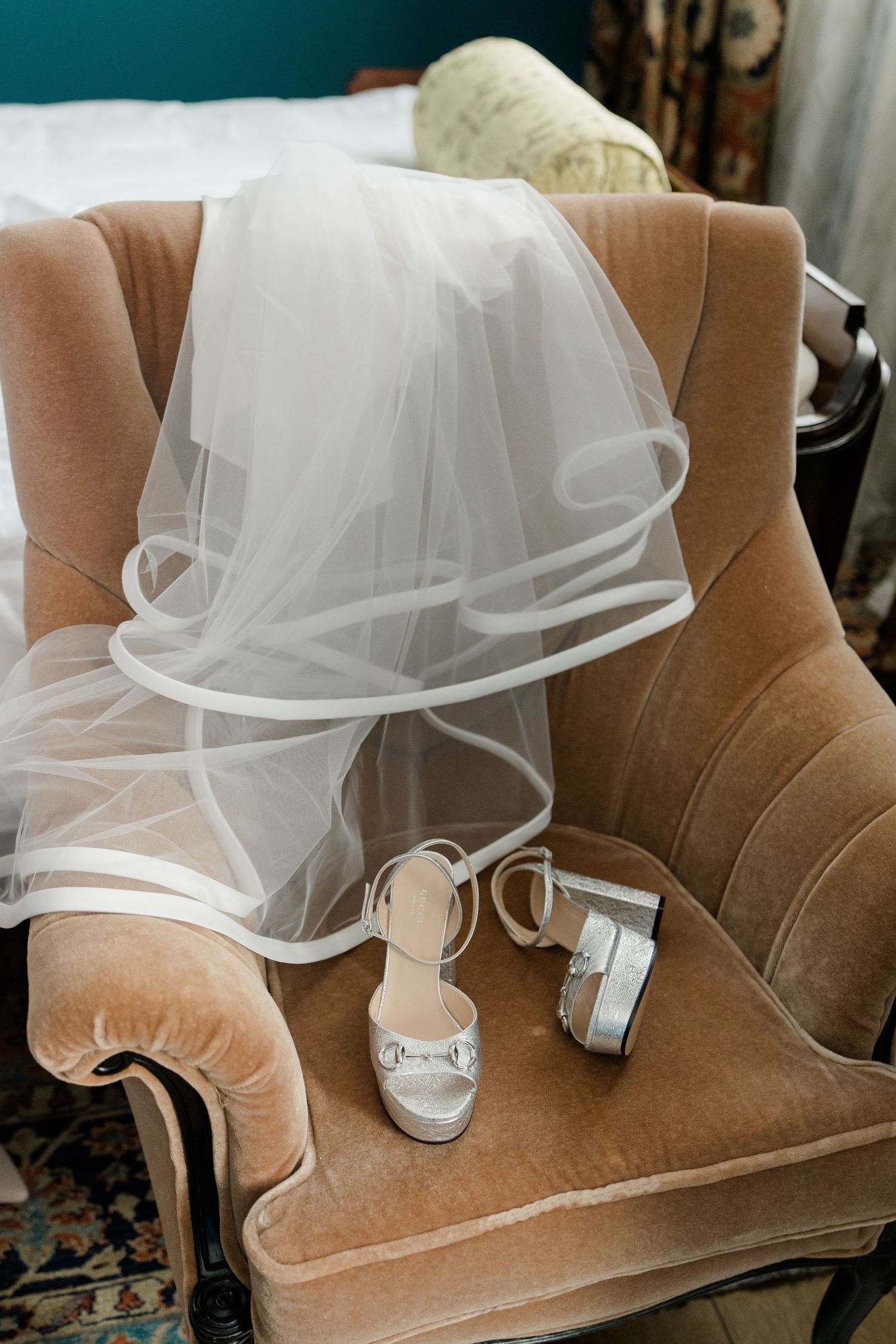 White bridal veil draped over velvet chair with silver wedding heels and small purse on seat.