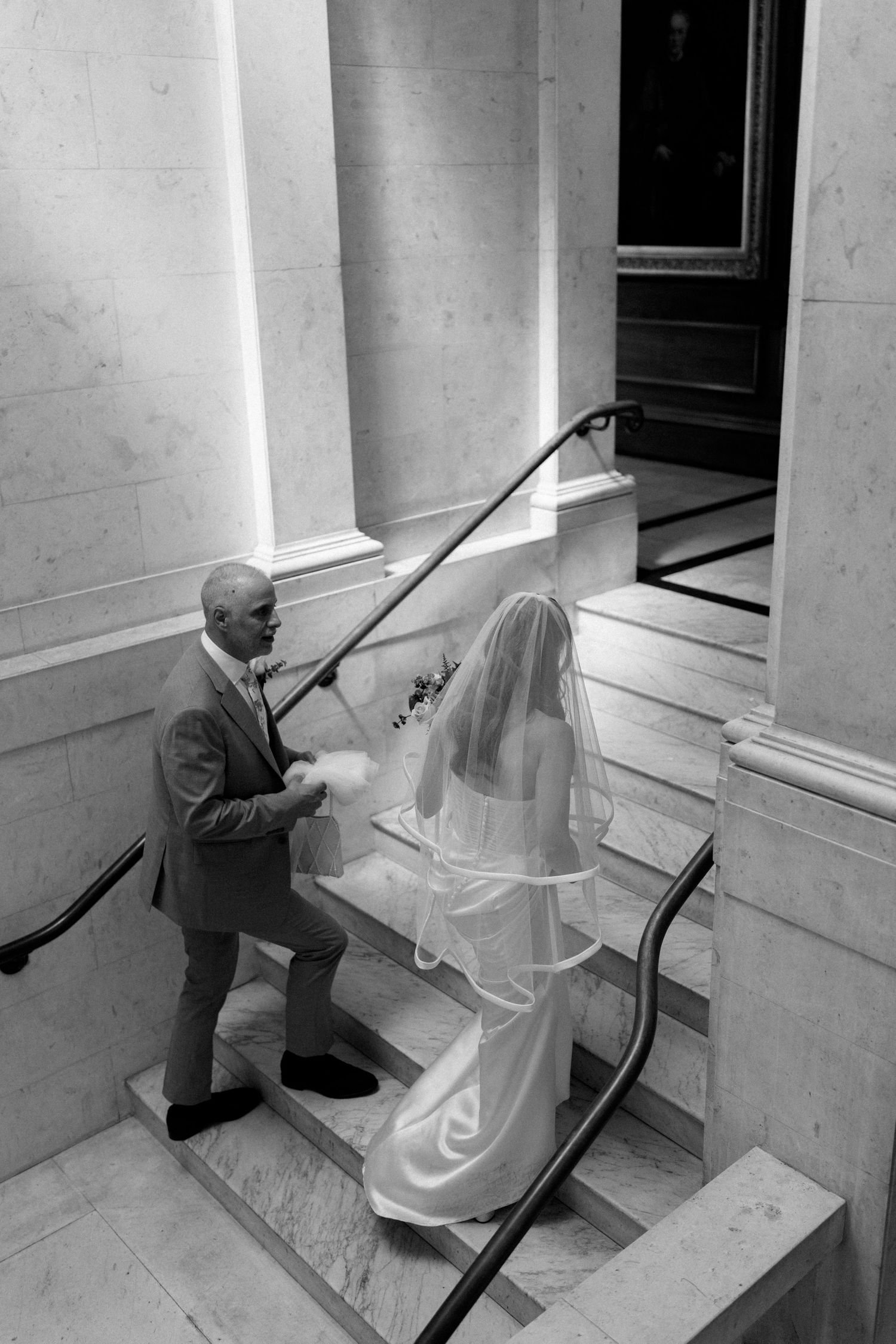 Black and white overhead view of groom assisting bride up marble staircase in grand venue.