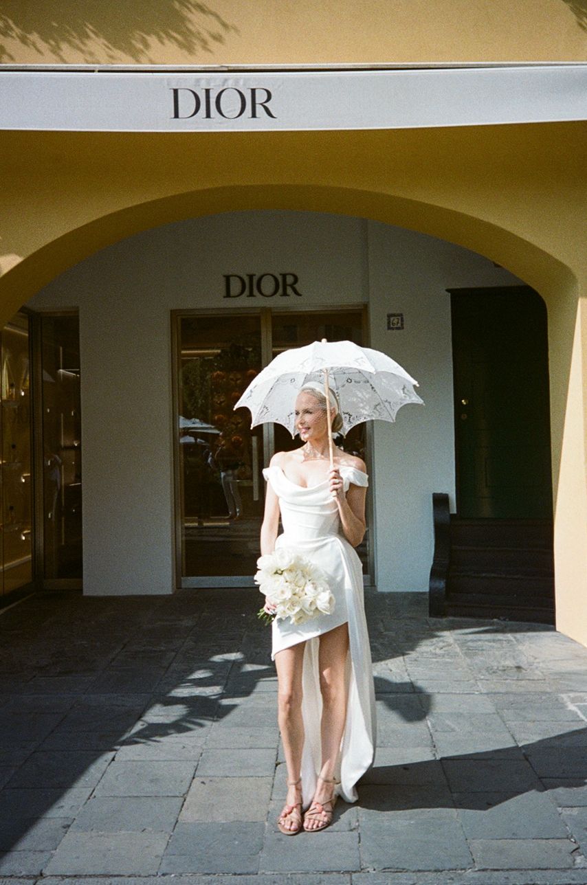 Bride in white mini dress holding lace parasol and bouquet outside Dior boutique.