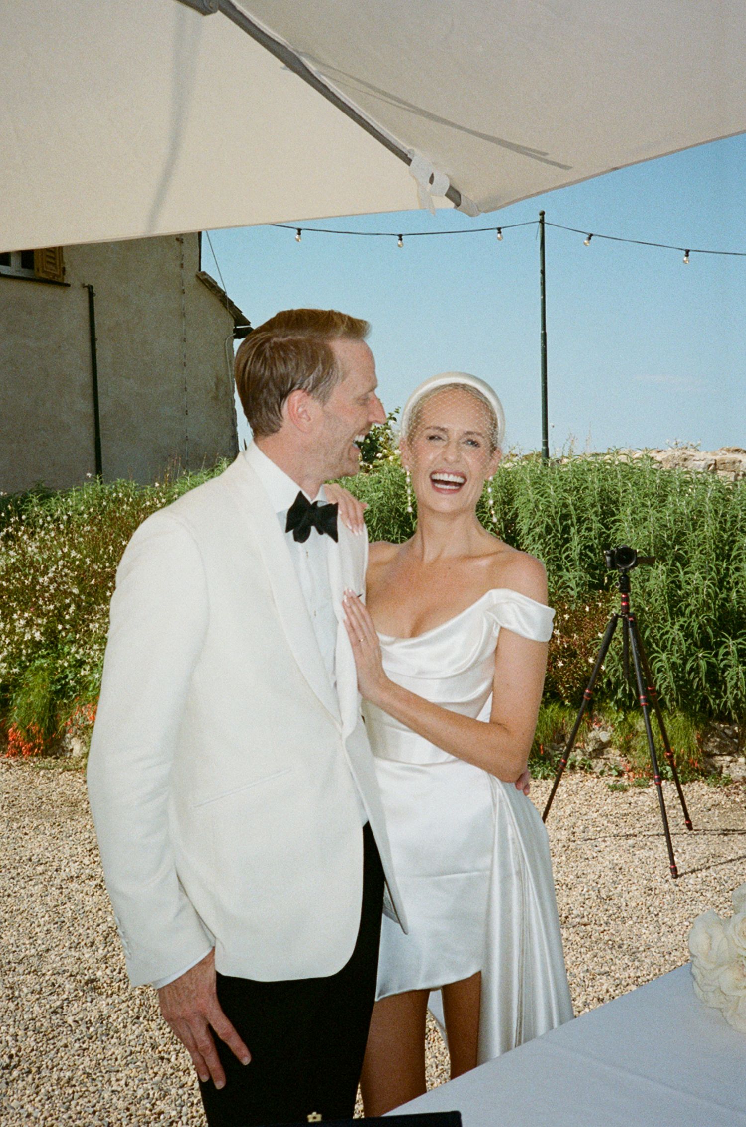 Laughing bride in off-shoulder white mini dress with groom in white tuxedo at sunny outdoor Italian wedding reception.