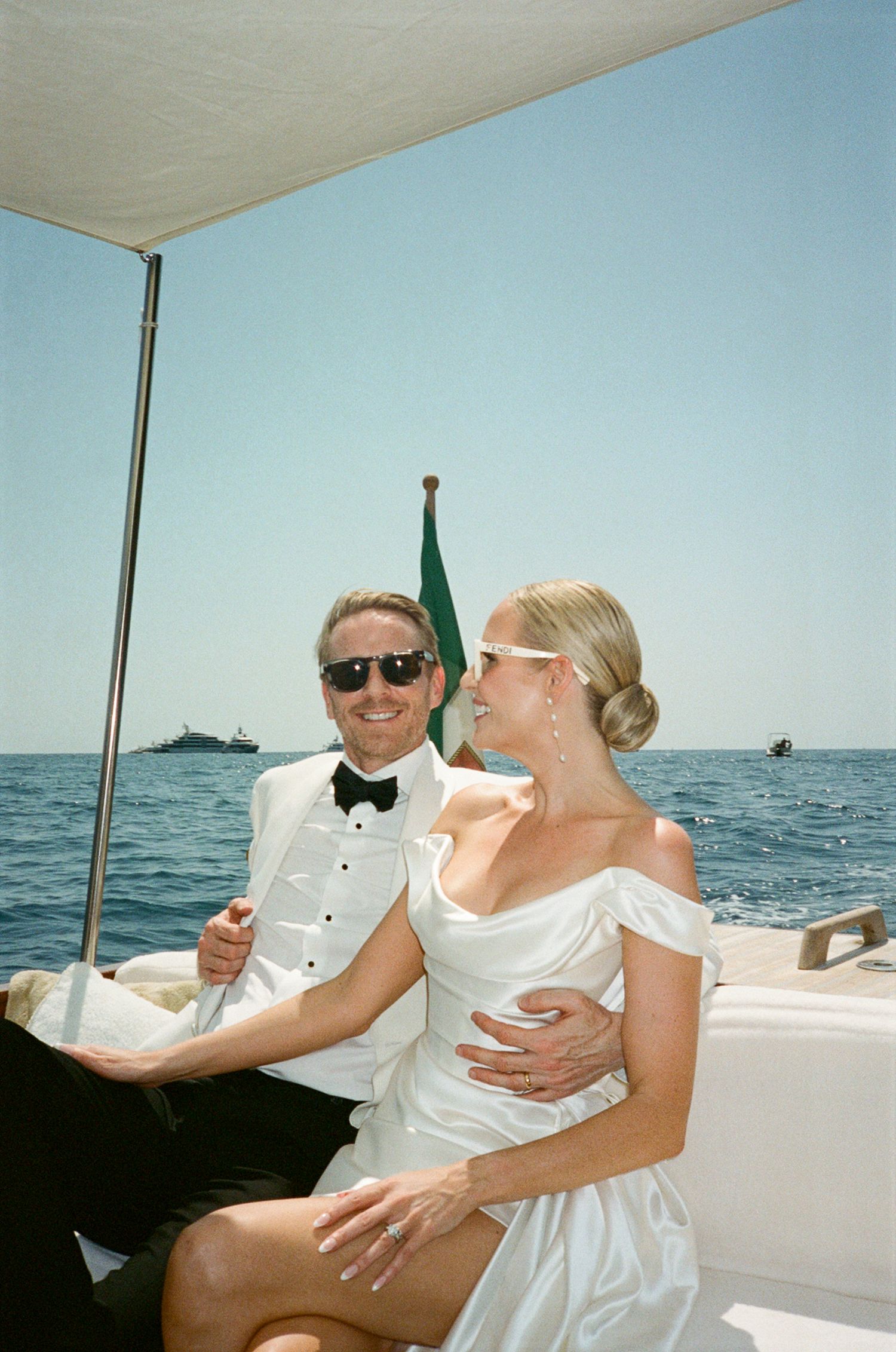 Elegant bride and groom in sunglasses embracing on a luxury boat with the Italian Riviera sea in the background.