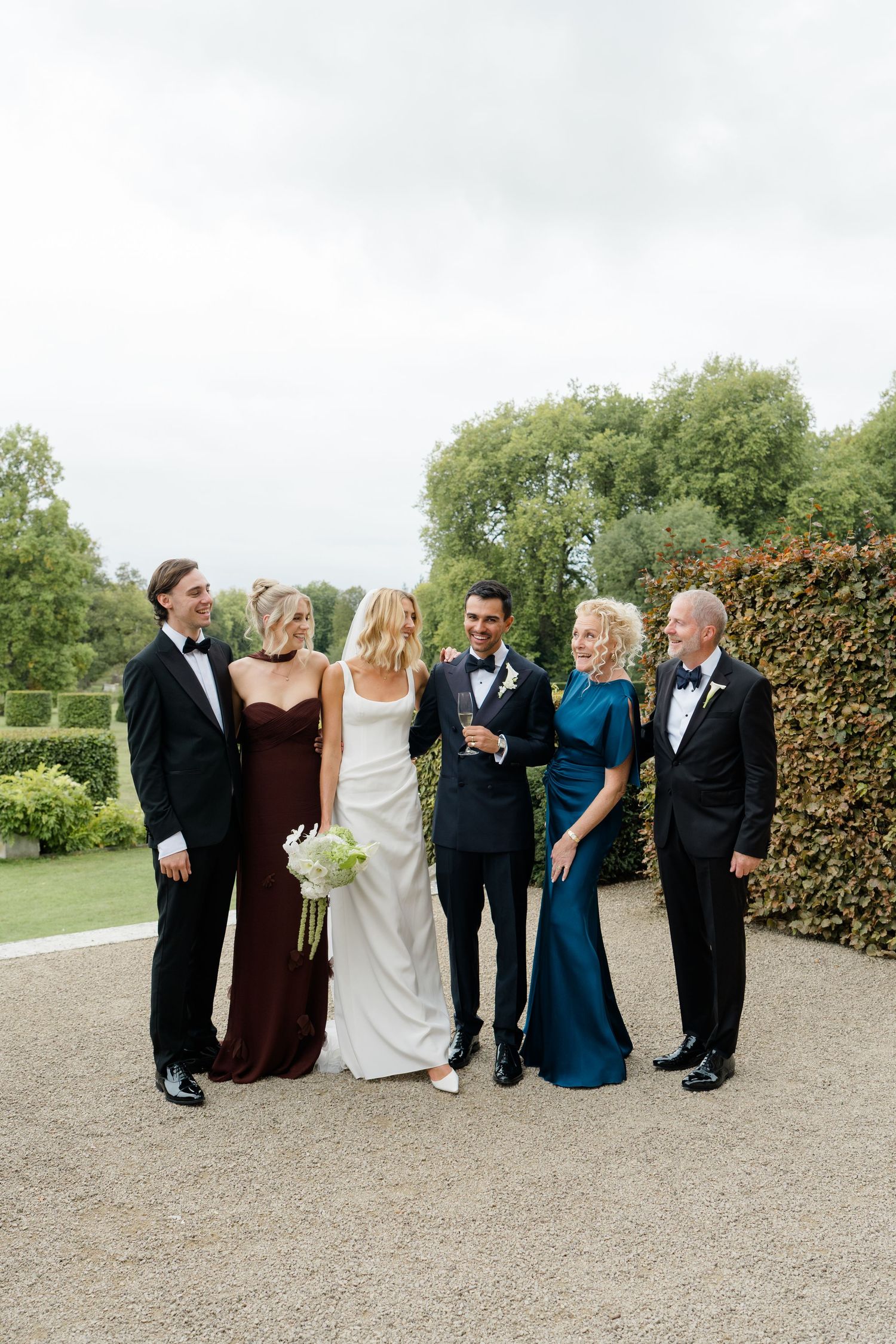Wedding party of six posing outdoors in formal attire on gravel path.