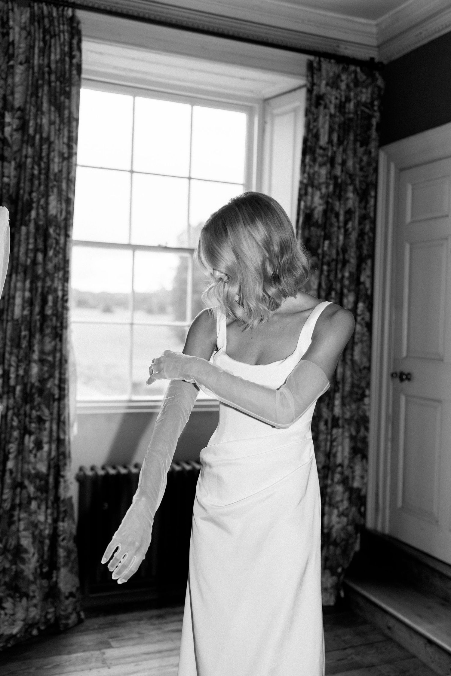 Bride in white gown putting on lace gloves by a window in black and white photo.