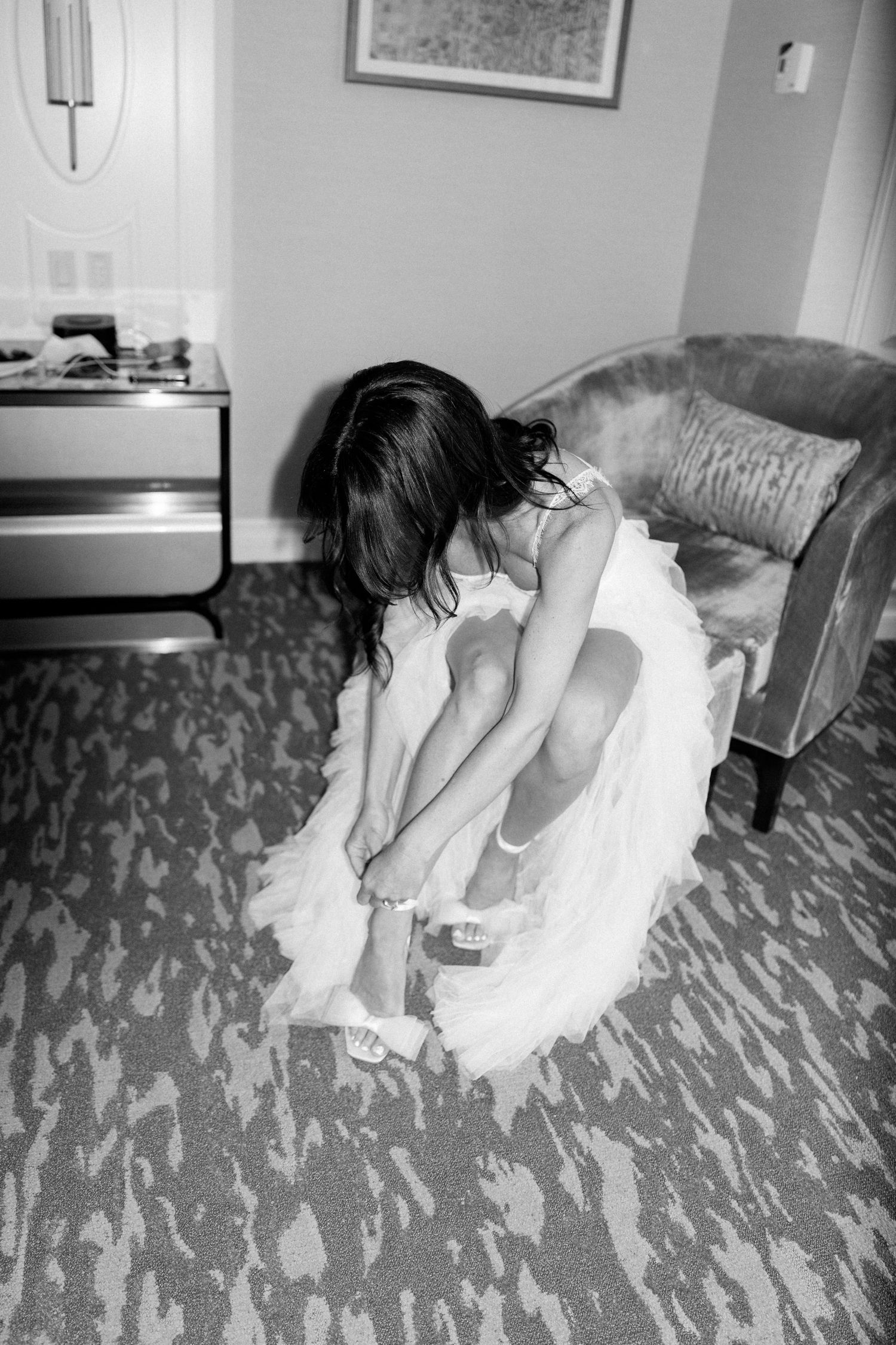 Bride in white dress putting on shoes while seated in preparation room.