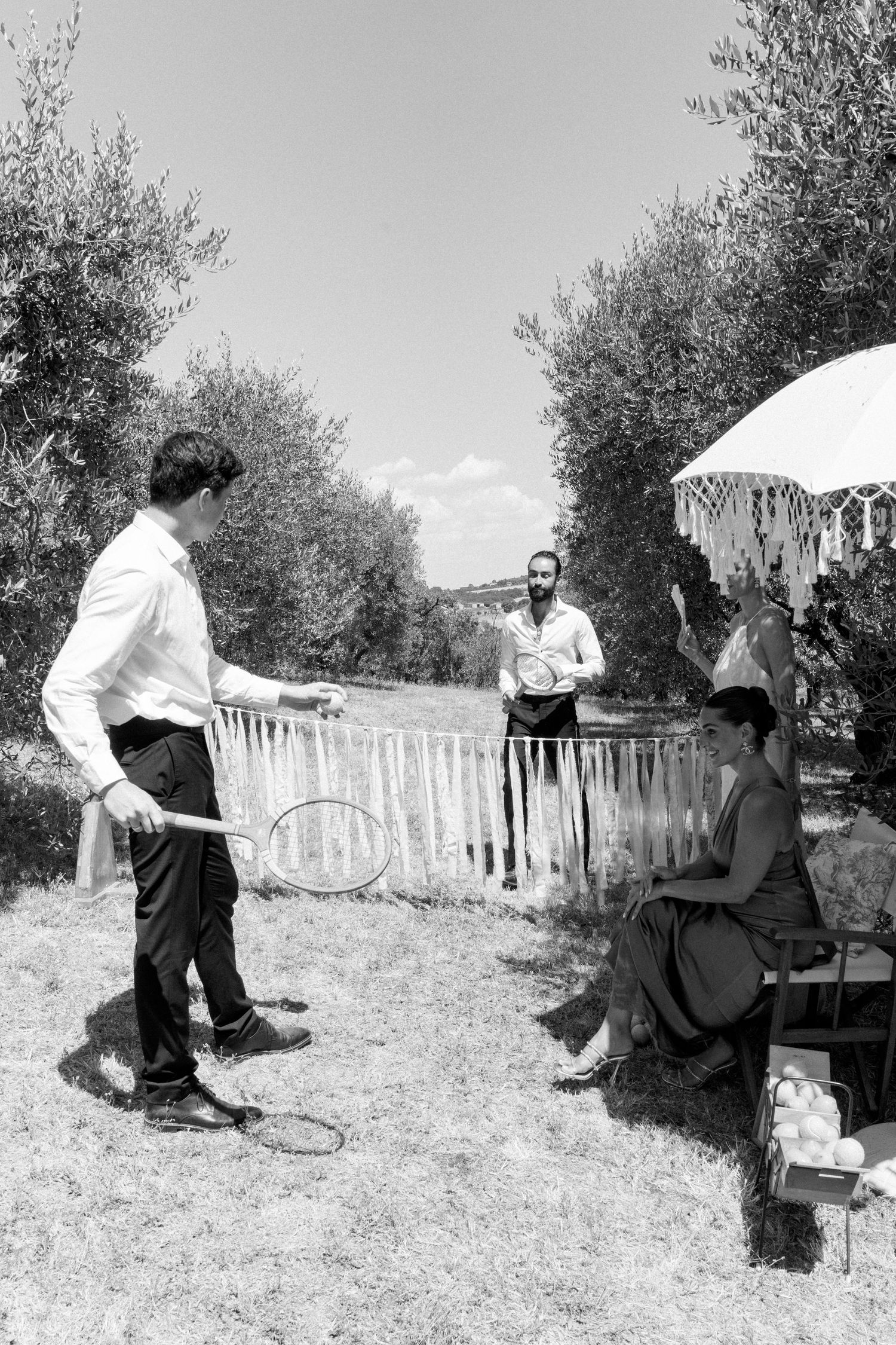 Black and white photo of guests socializing outdoors in an olive grove with white fencing.
