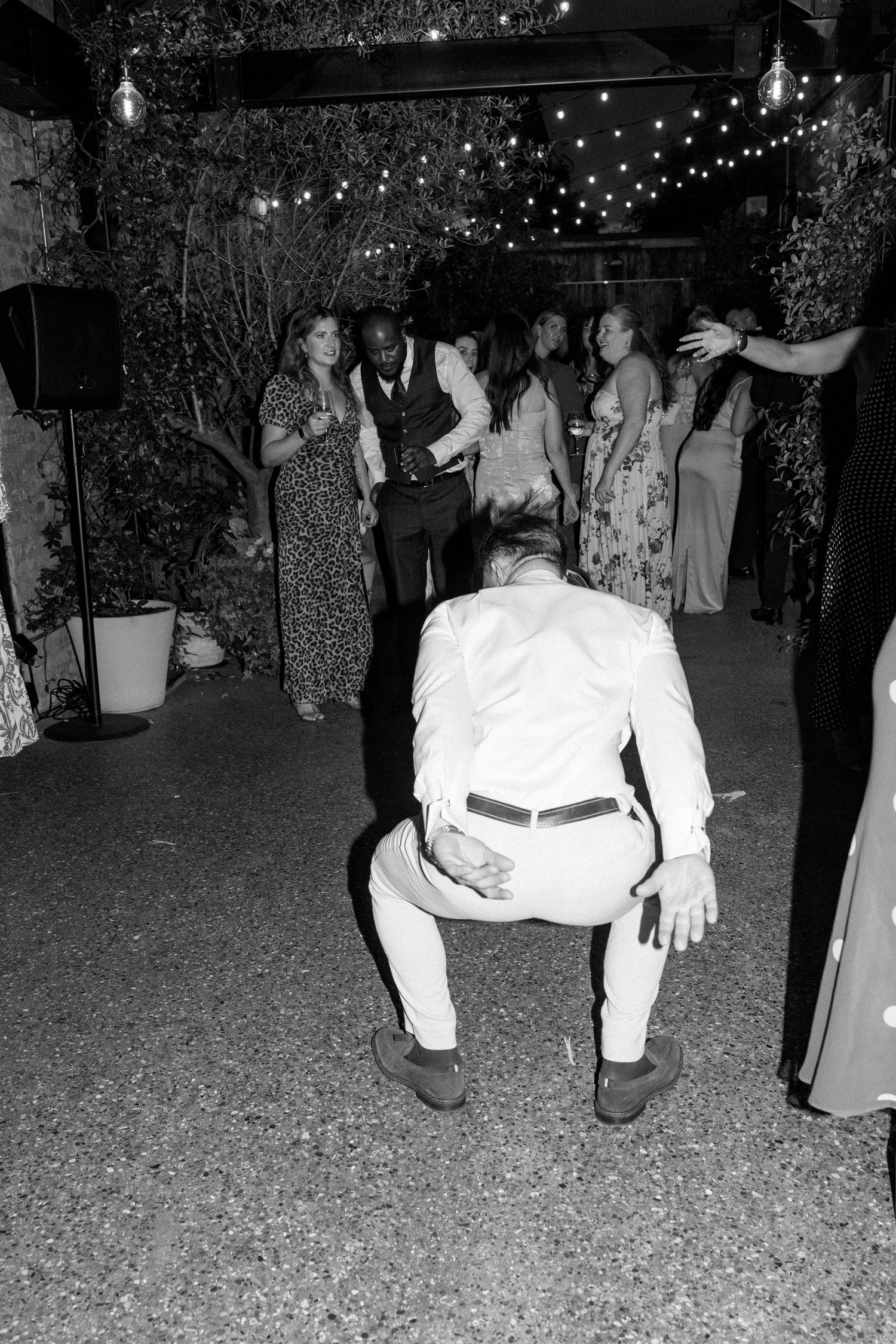 Black and white photo of wedding guests dancing on gravel floor under string lights.