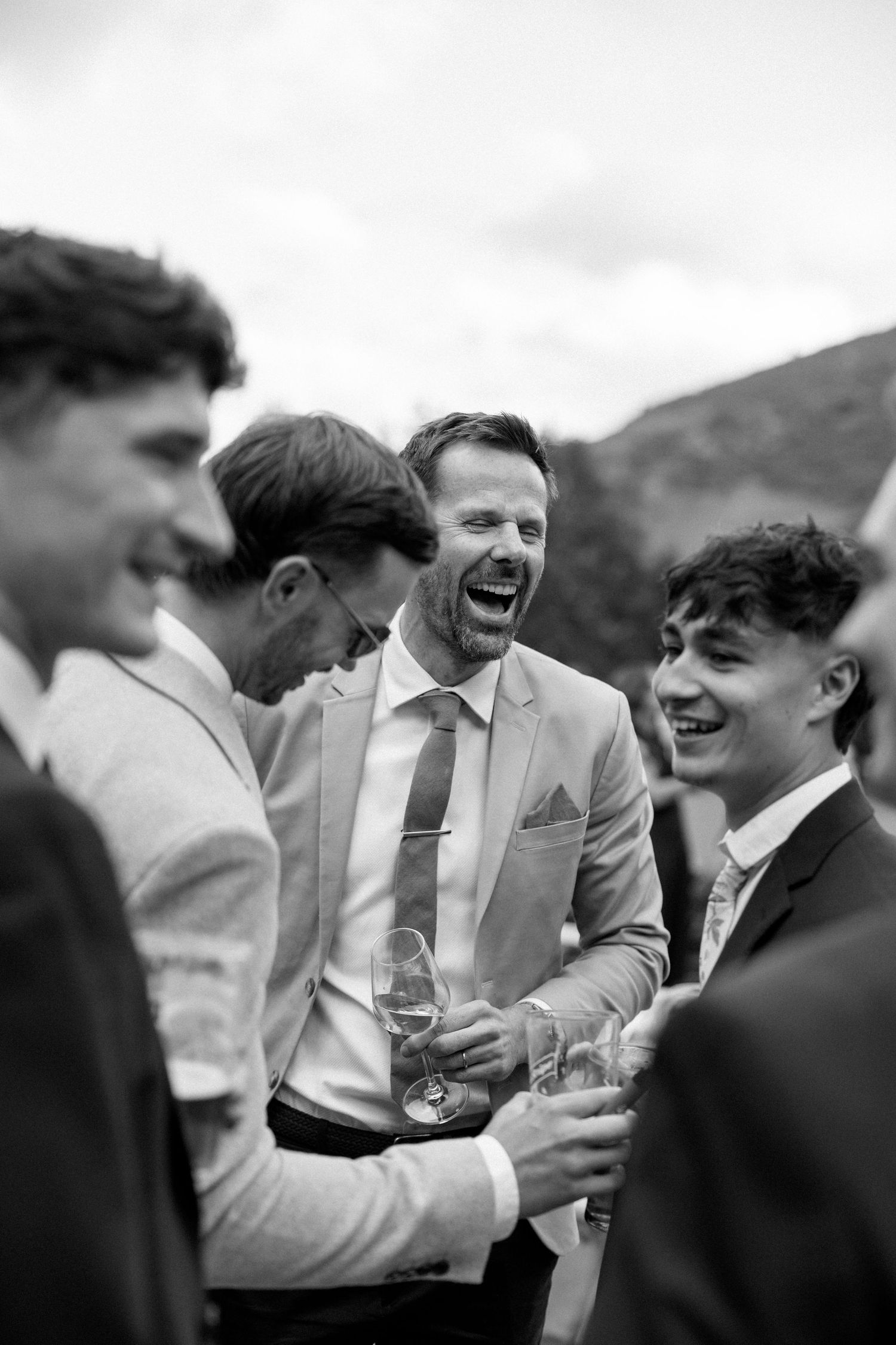 Groomsmen in light suits share drinks and laughter during outdoor wedding festivities.
