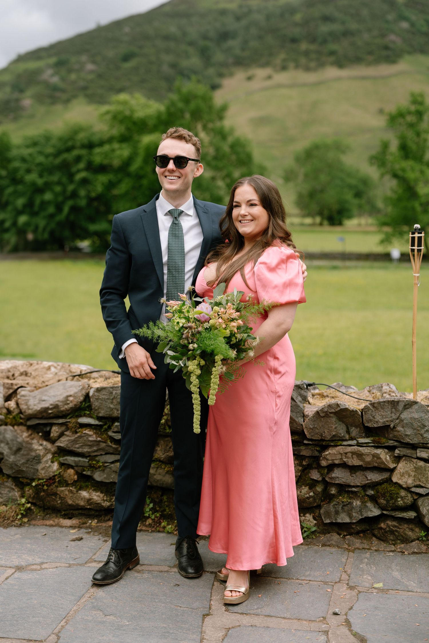 Wedding couple posing against stone wall with green hills in background.