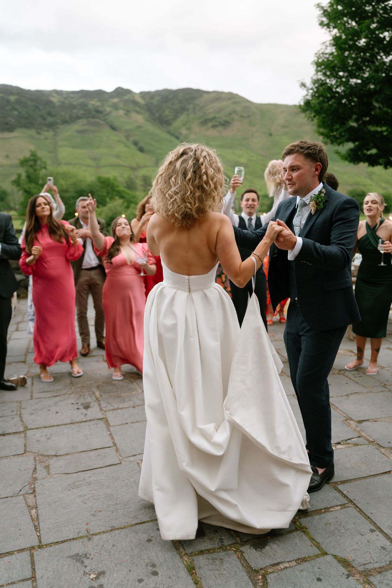 Wedding celebration with guests dancing in coral dresses against a mountainous backdrop.