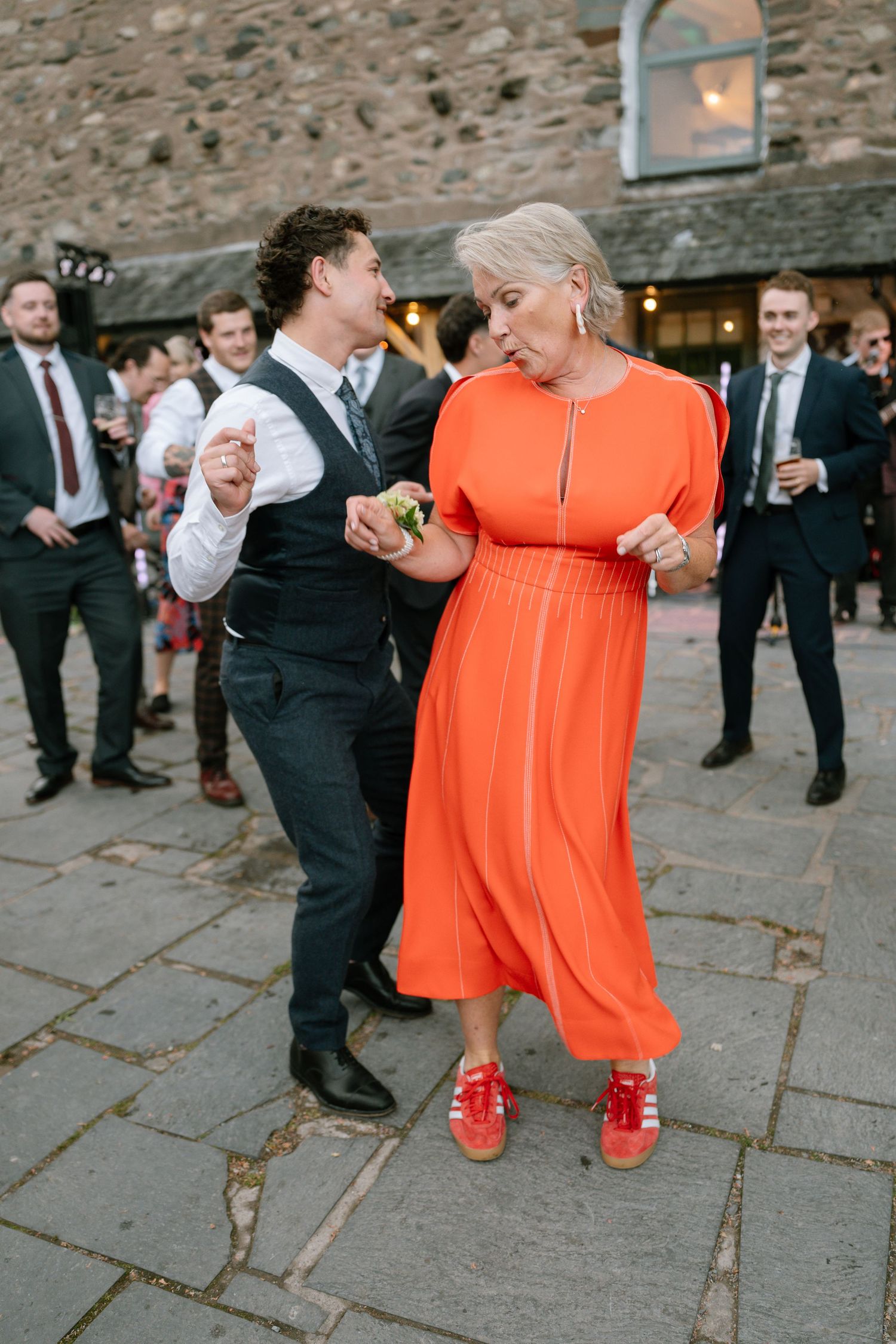 People in formal attire and bright orange dresses dance on a stone patio during a celebration.