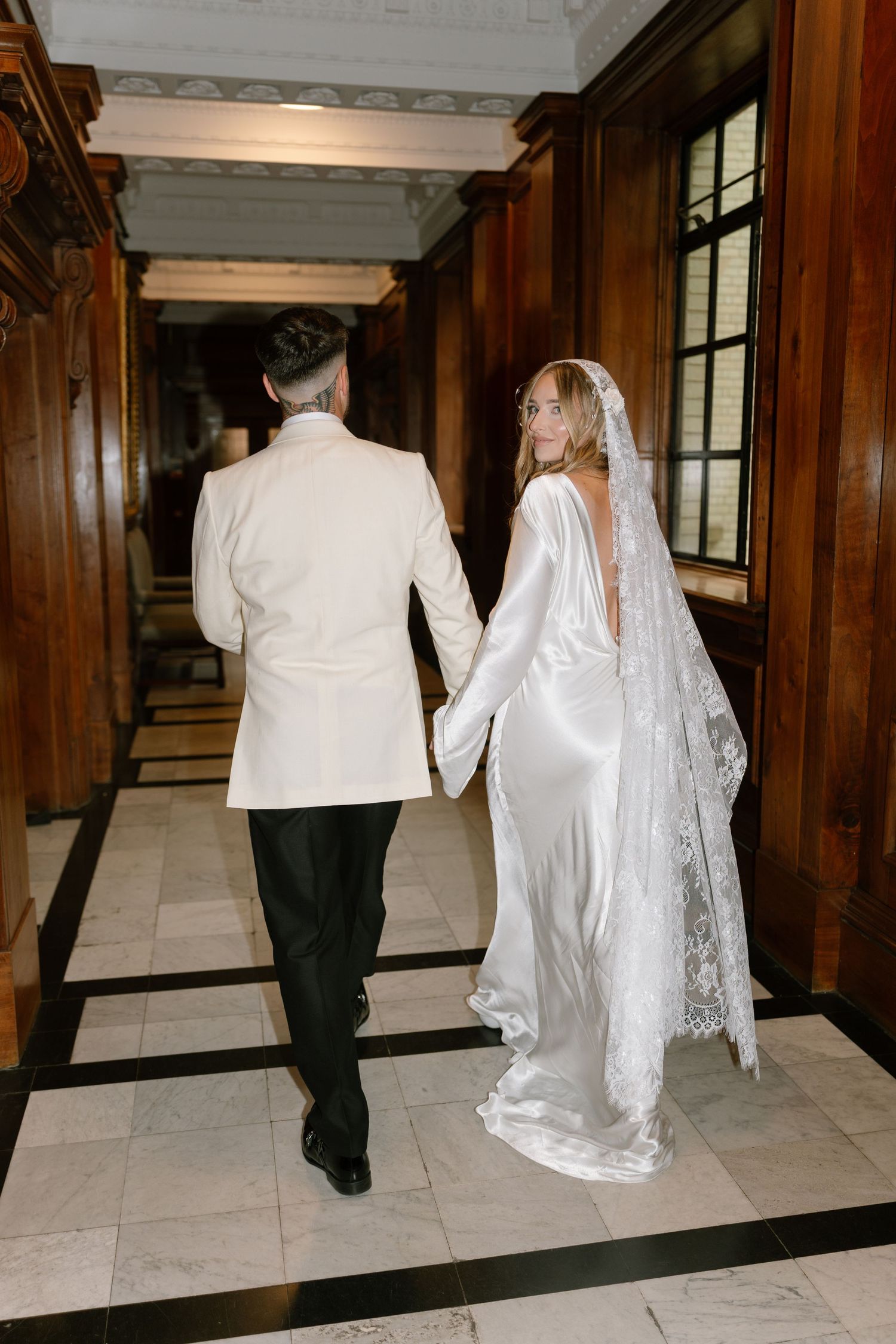 Wedding couple walking down a grand hallway with wood paneling and marble floors.