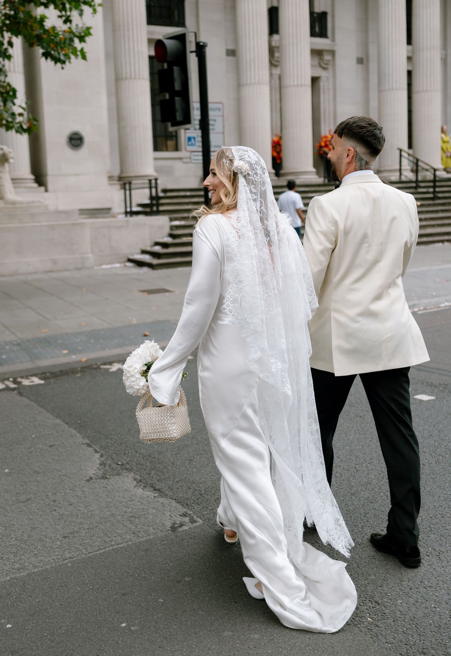 A bride in a sleek white gown and veil walks alongside a person in a white jacket on a city street.