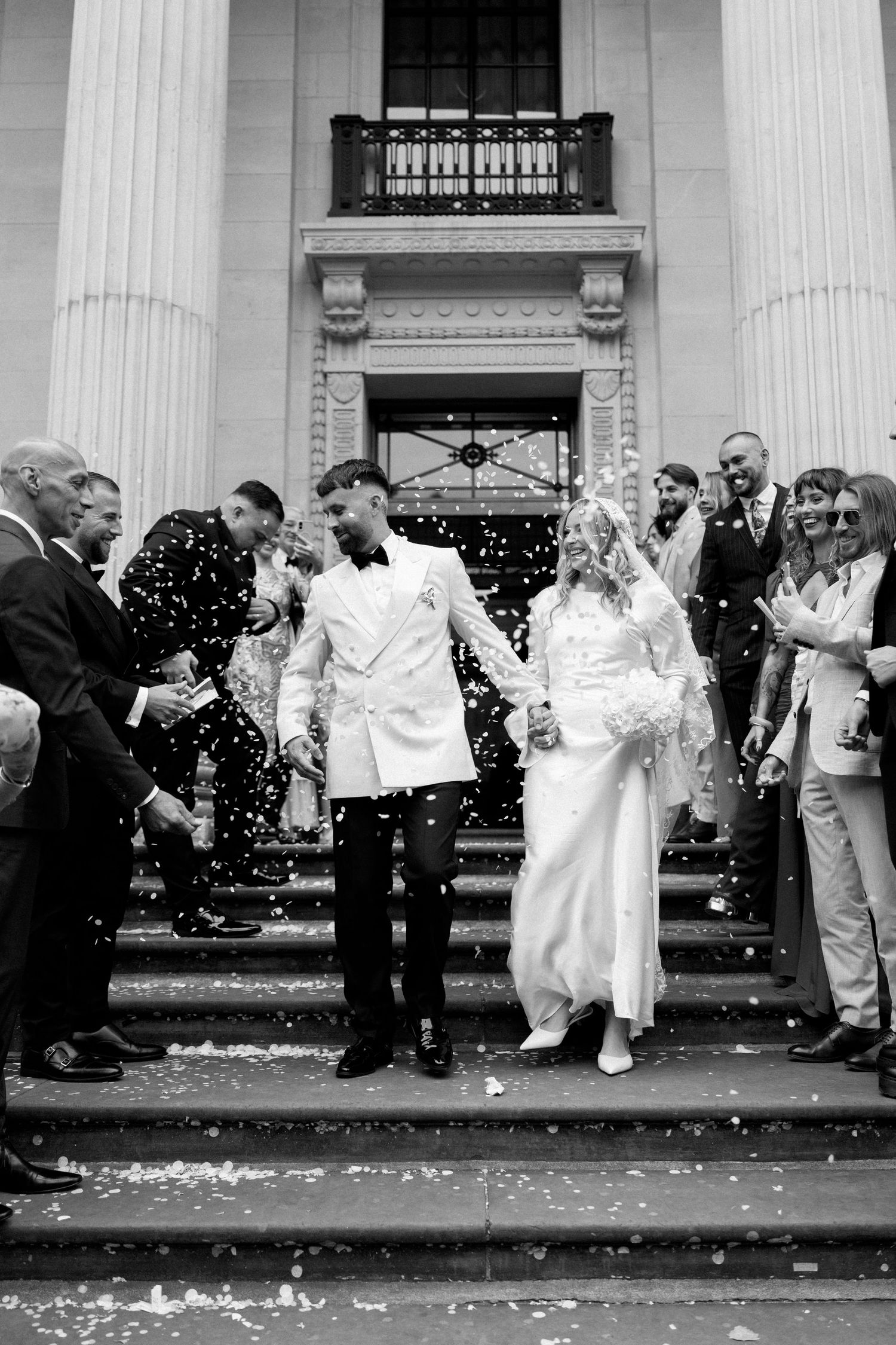 Wedding party and guests celebrate with confetti outside a grand building with white columns.