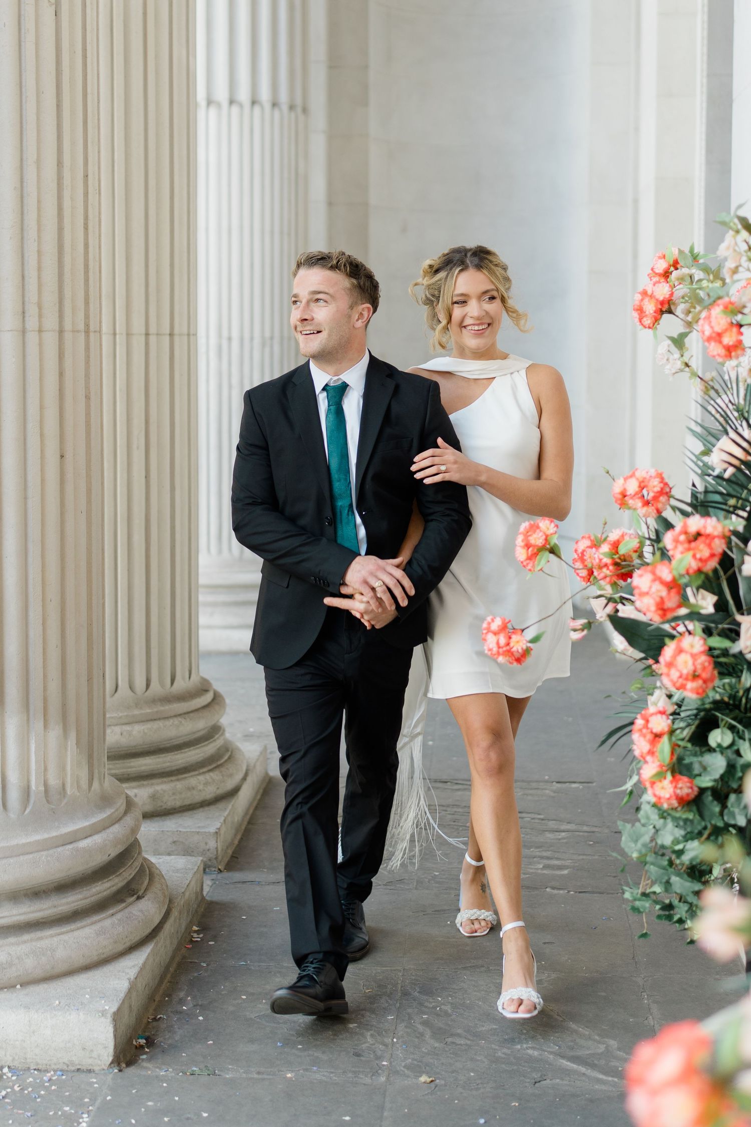 A couple poses playfully between white columns with coral-colored flowers in the foreground.