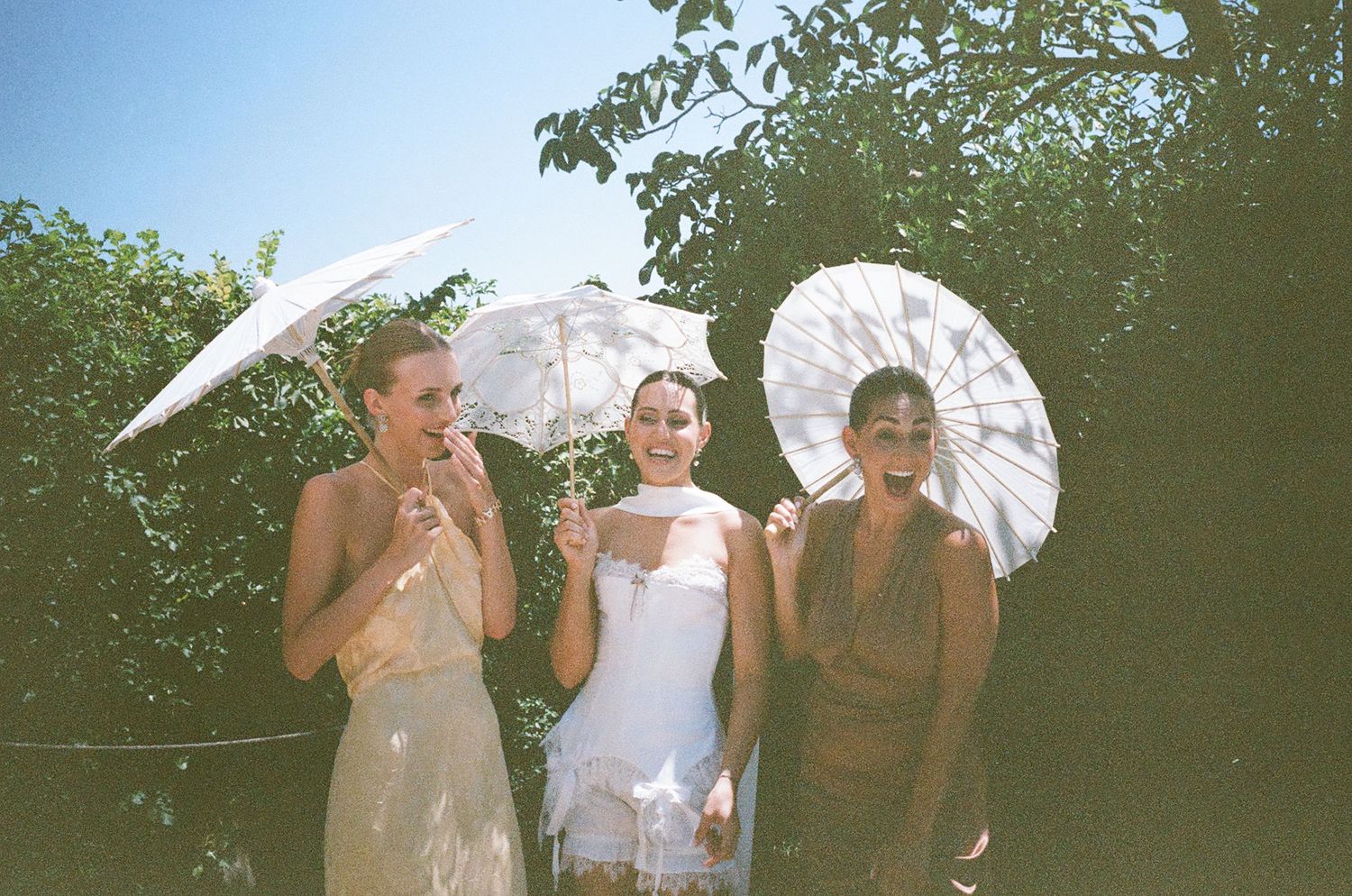 Three people in vintage-style clothing pose with parasols in a garden setting during what appears to be a summer photoshoot.