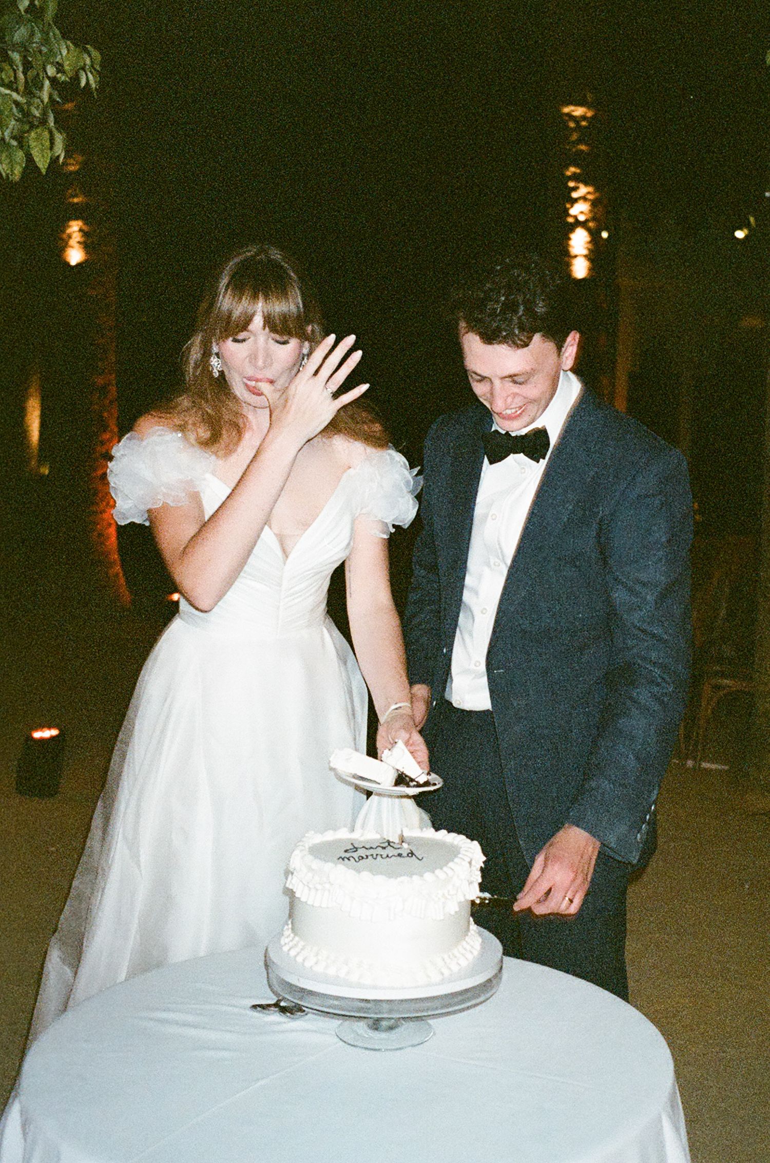 A couple at their wedding reception cutting into a white tiered cake together in a dimly lit venue.