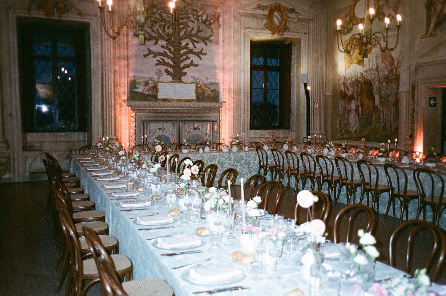 Elegant banquet hall setup with long tables, white linens, and ornate chandeliers in a luxurious ballroom setting.