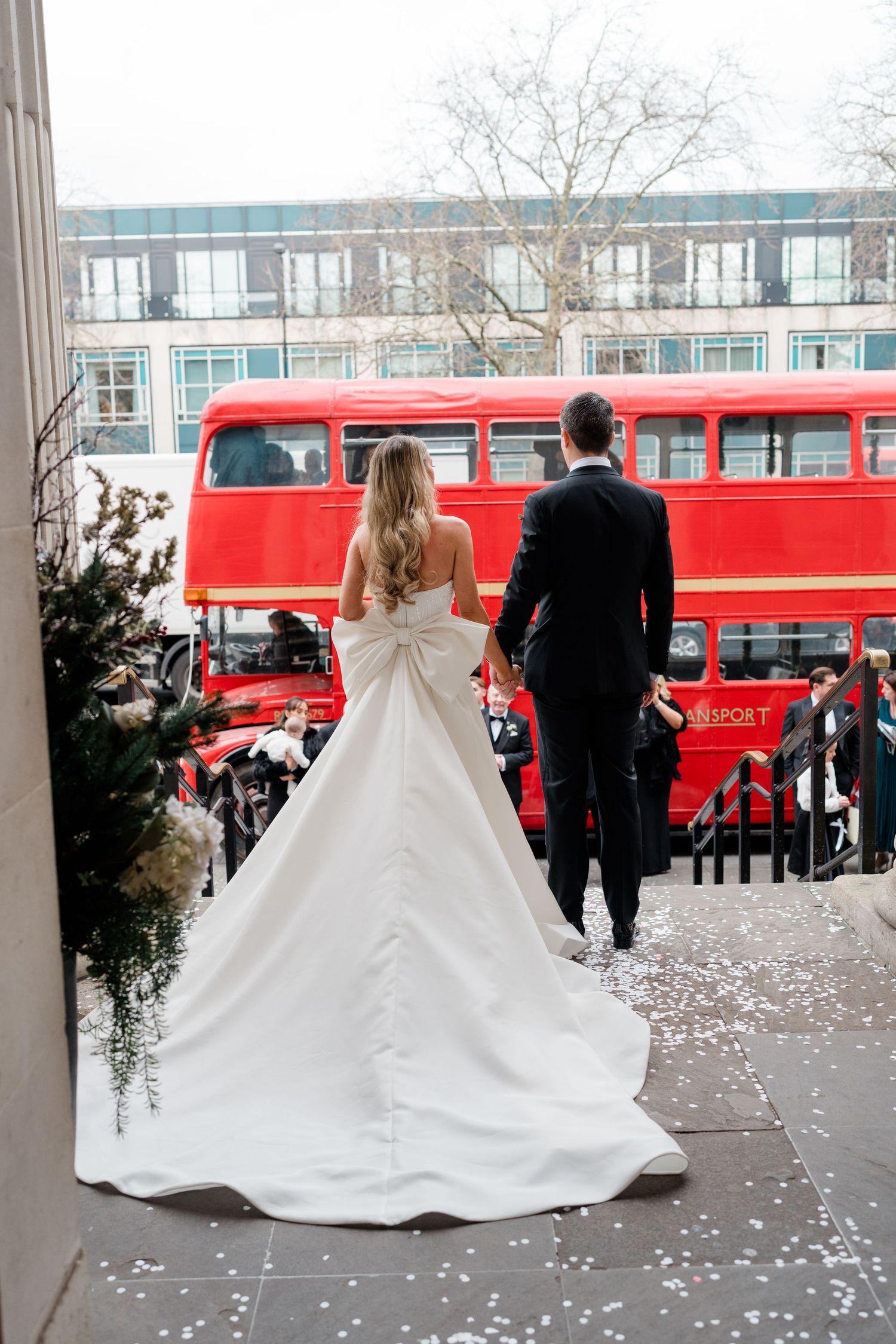 Wedding couple poses in front of red London double-decker buses with scattered confetti.