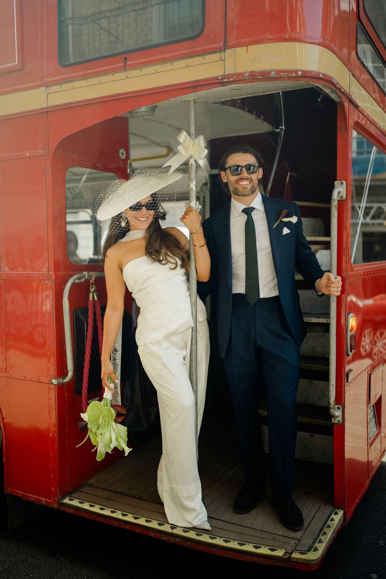 Stylish bride and groom in sunglasses pose on iconic red London double-decker bus on wedding day.