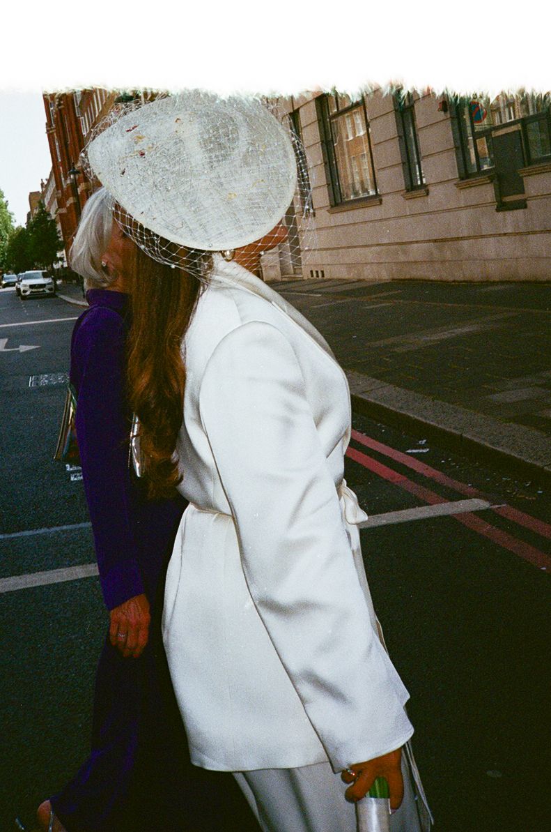 Woman in white coat and fascinator hat walks city street beside woman in purple dress.