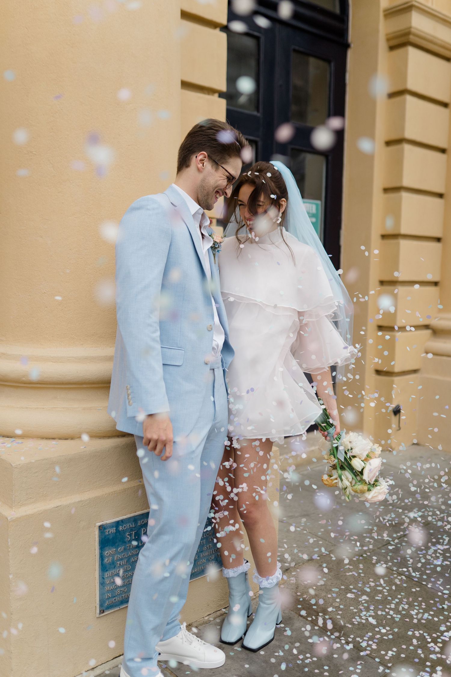Newlywed couple sharing a romantic moment outdoors, surrounded by falling confetti, bride in short white dress and groom in light blue suit.