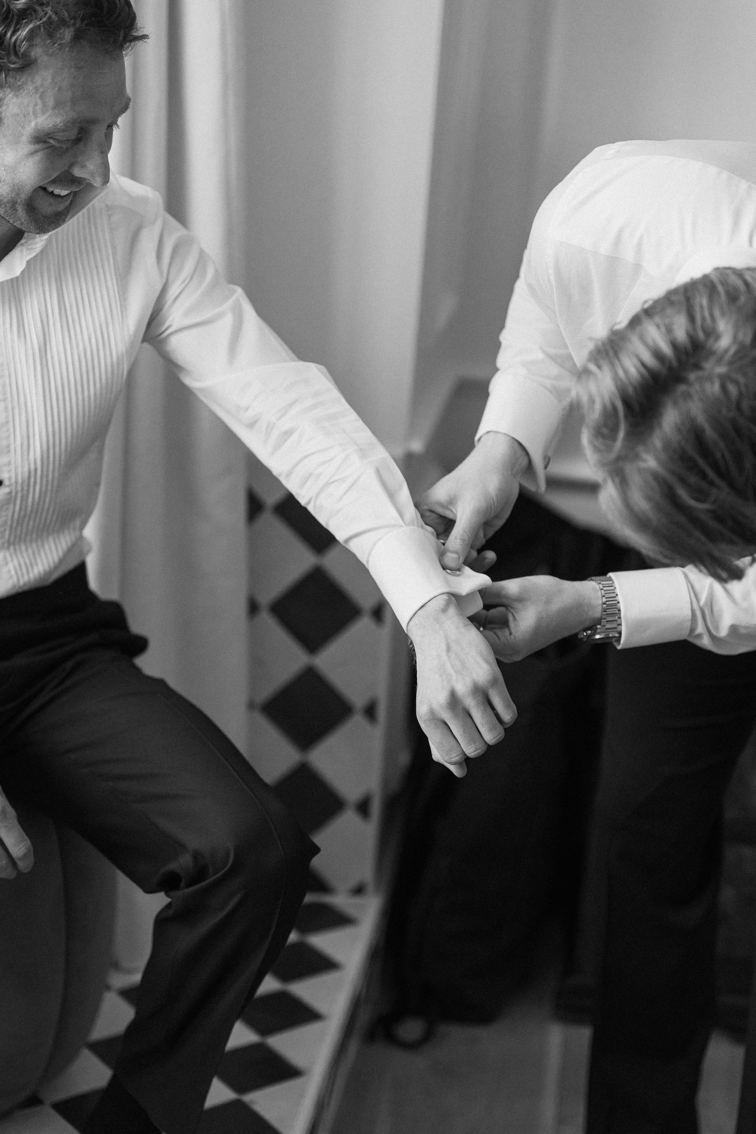 Black and white photo of groomsman helping groom fasten cufflinks on black and white checkered floor.