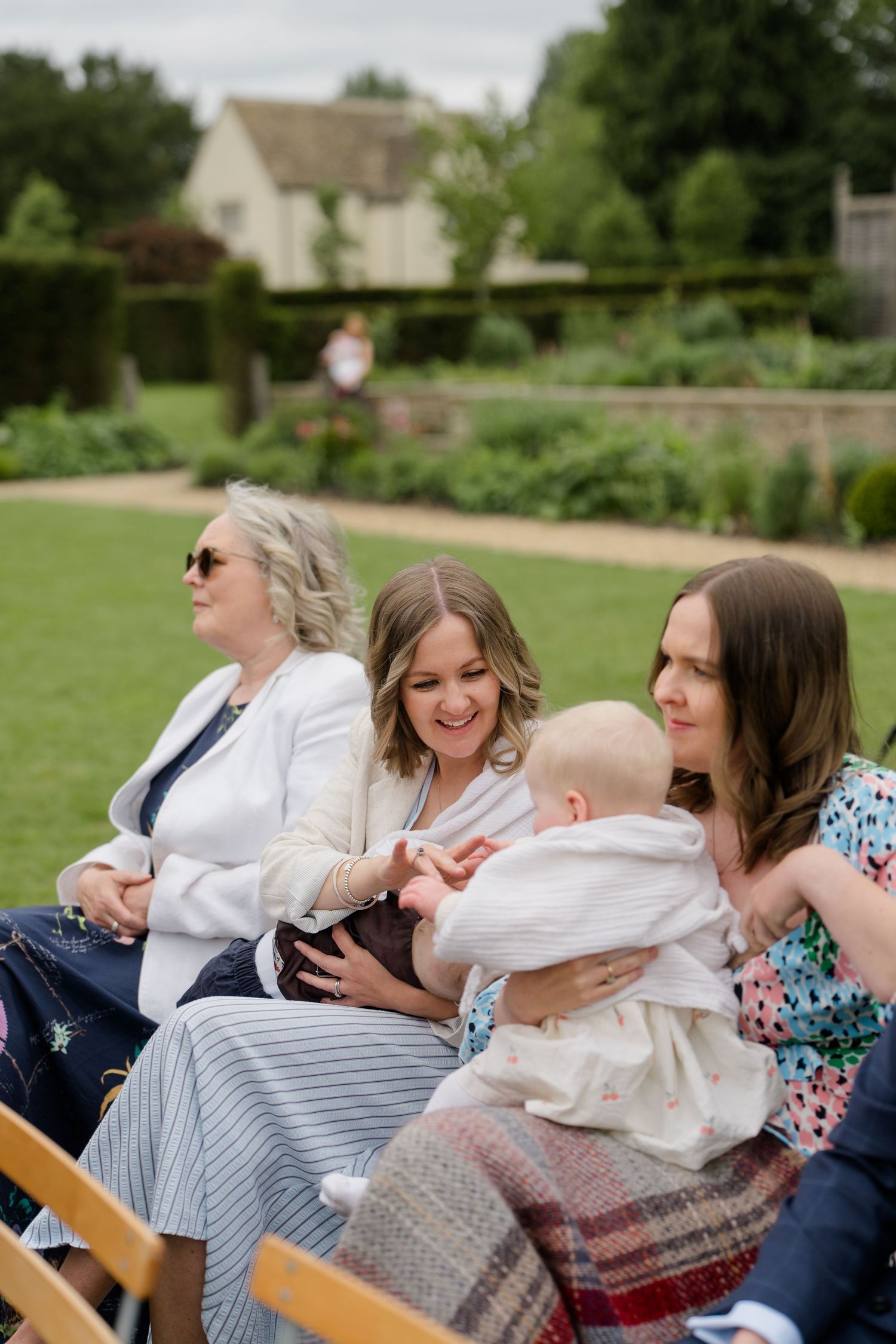 Women guests smile and hold a baby during an outdoor garden wedding ceremony.
