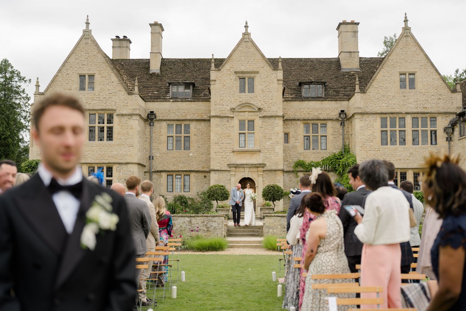 Outdoor wedding ceremony on lawn of grand English manor house with seated guests.