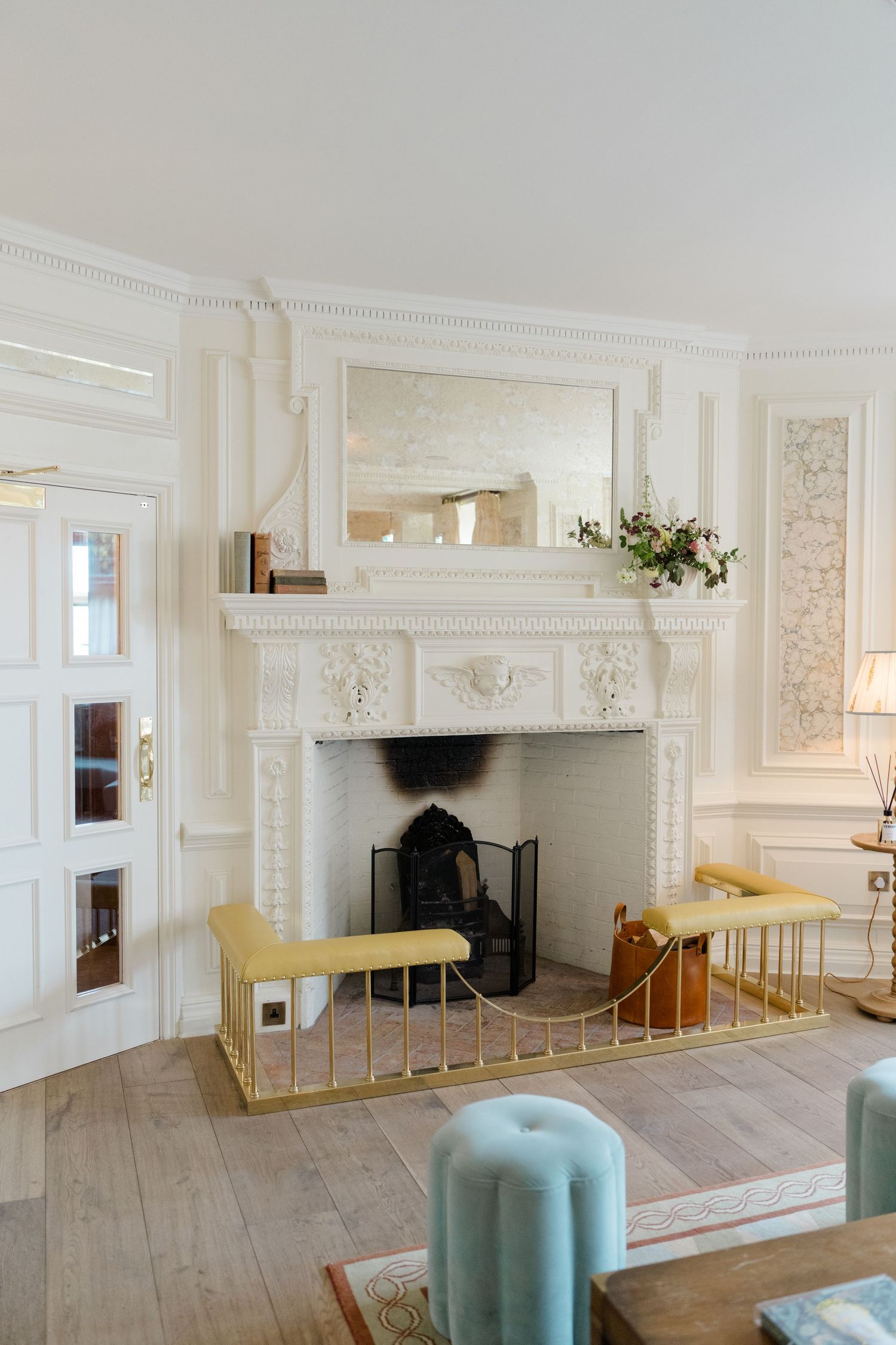 Ornate white carved fireplace with gold fender, mirror above mantel, and light wood floors in a classic interior.