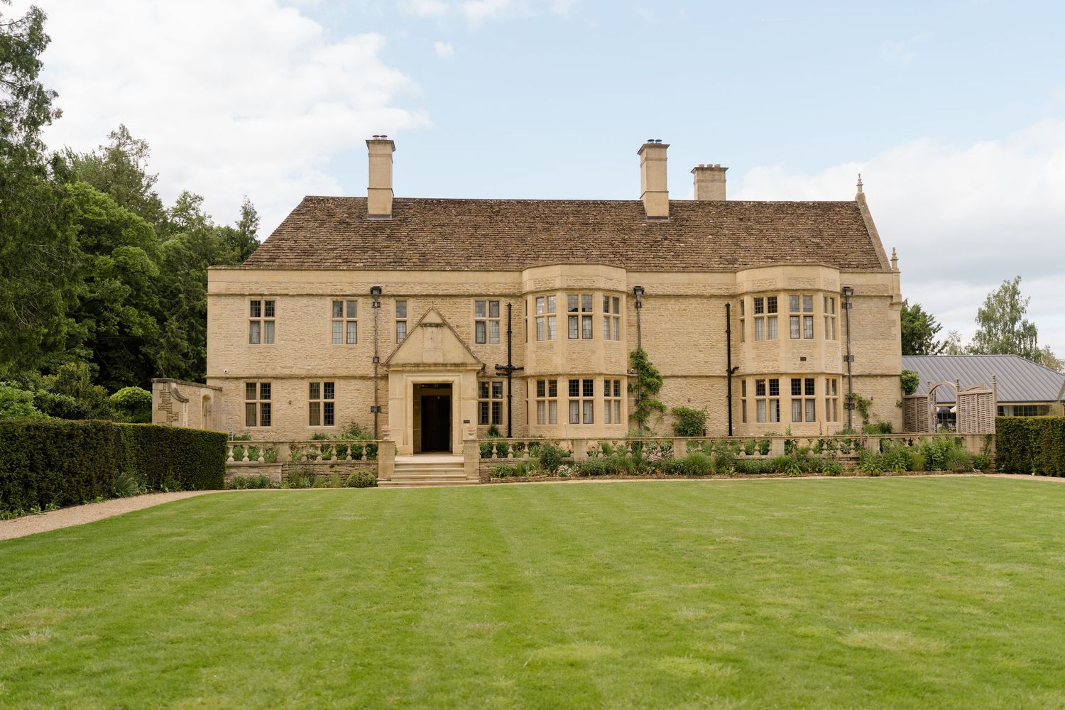 Grand Cotswold stone manor house with manicured lawn, mullioned windows, and symmetrical facade surrounded by mature hedges.