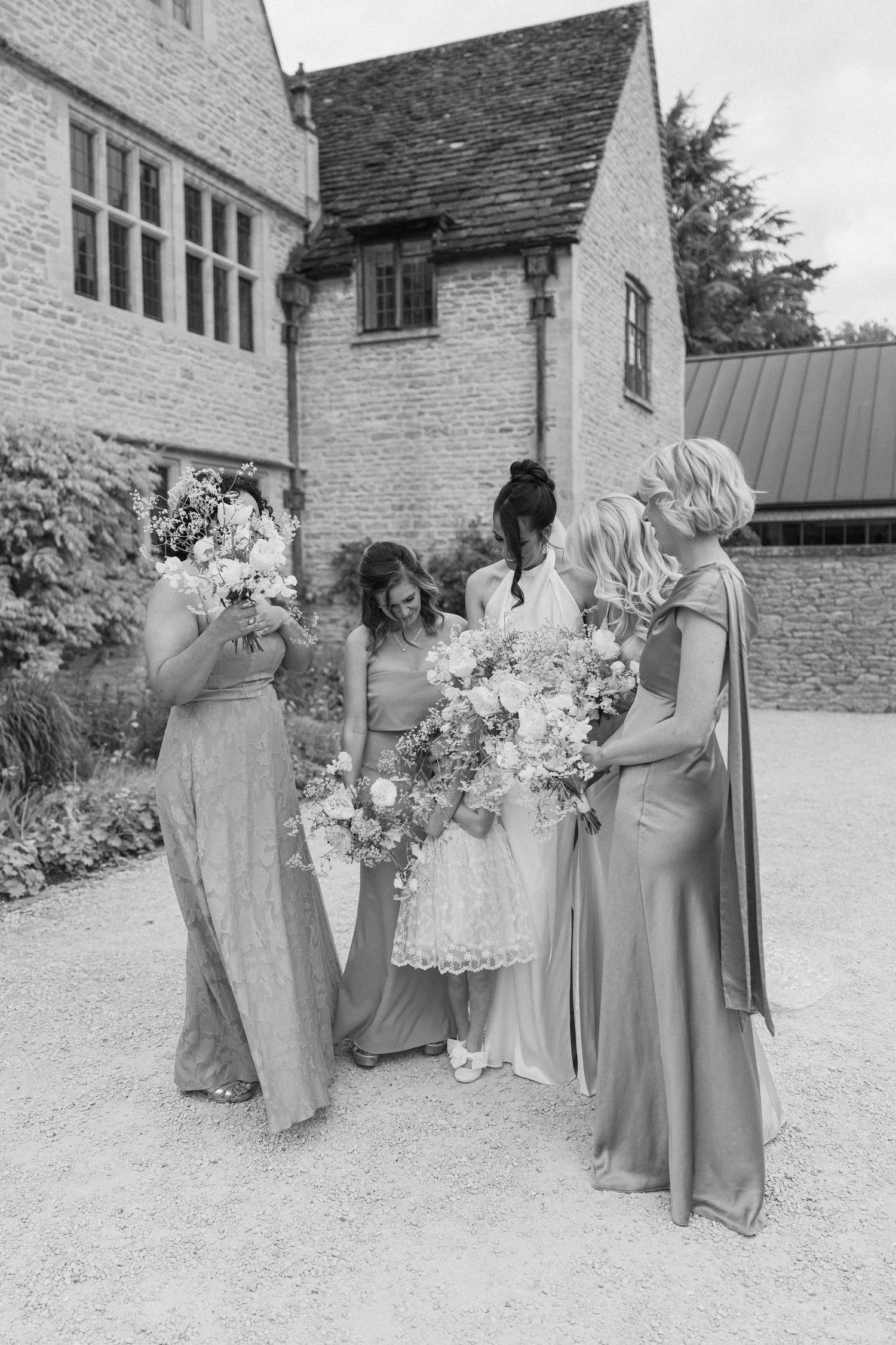 Black and white photo of bridesmaids in satin gowns admiring floral bouquets outside stone manor venue.