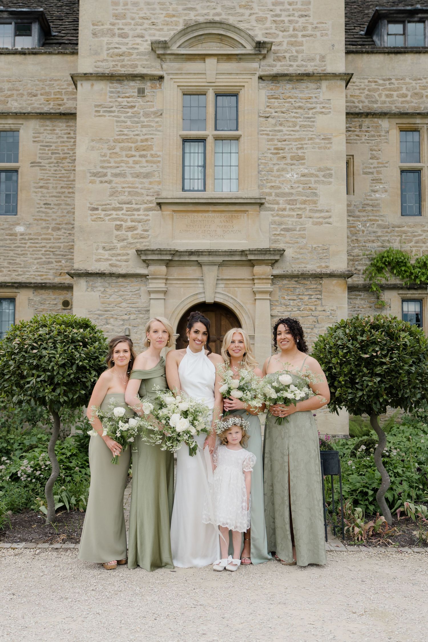 Bride poses with four bridesmaids in sage green gowns and flower girl outside stone manor.