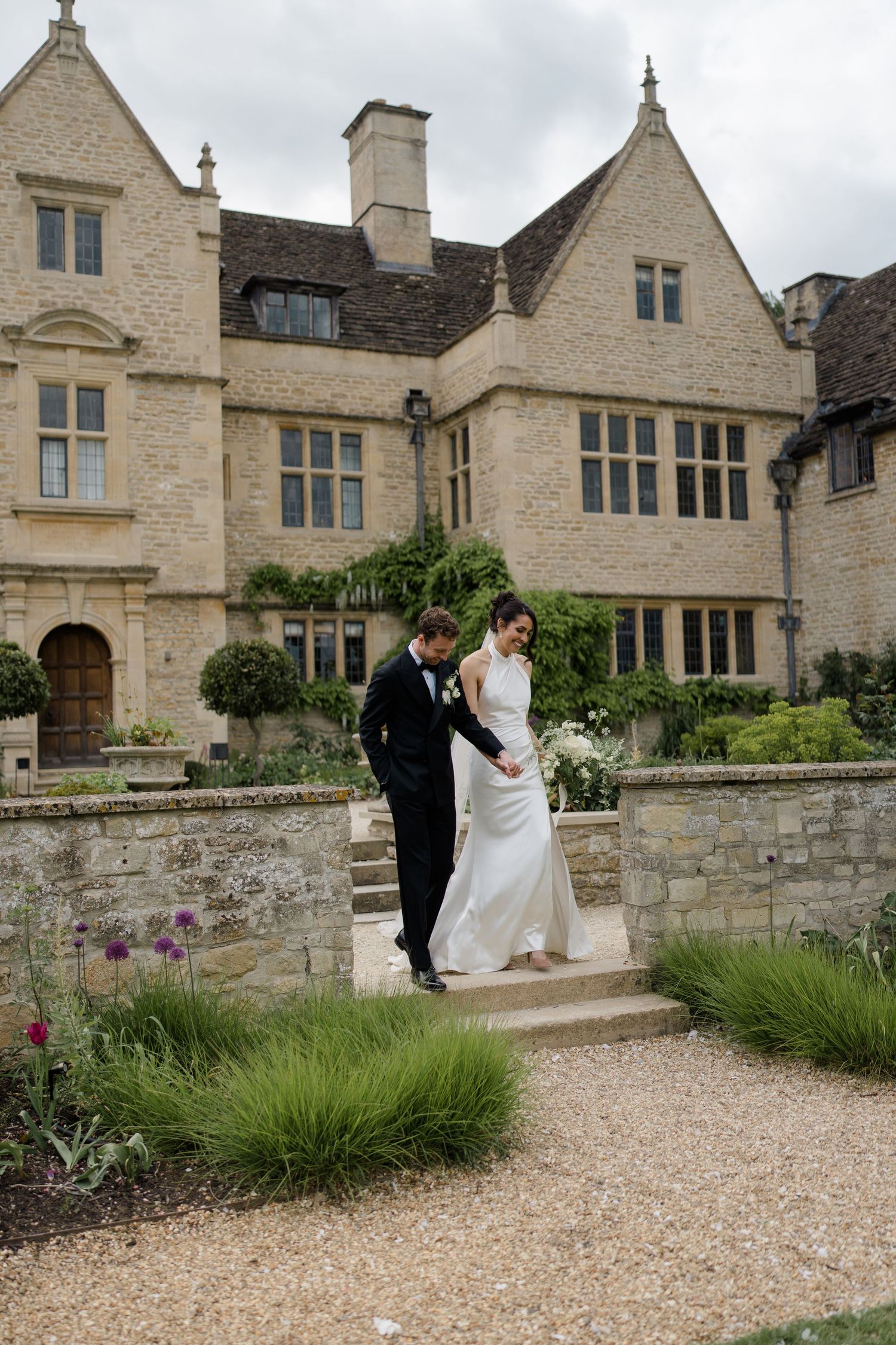 Groom helps bride descend garden steps at elegant stone manor house wedding venue on cloudy day.