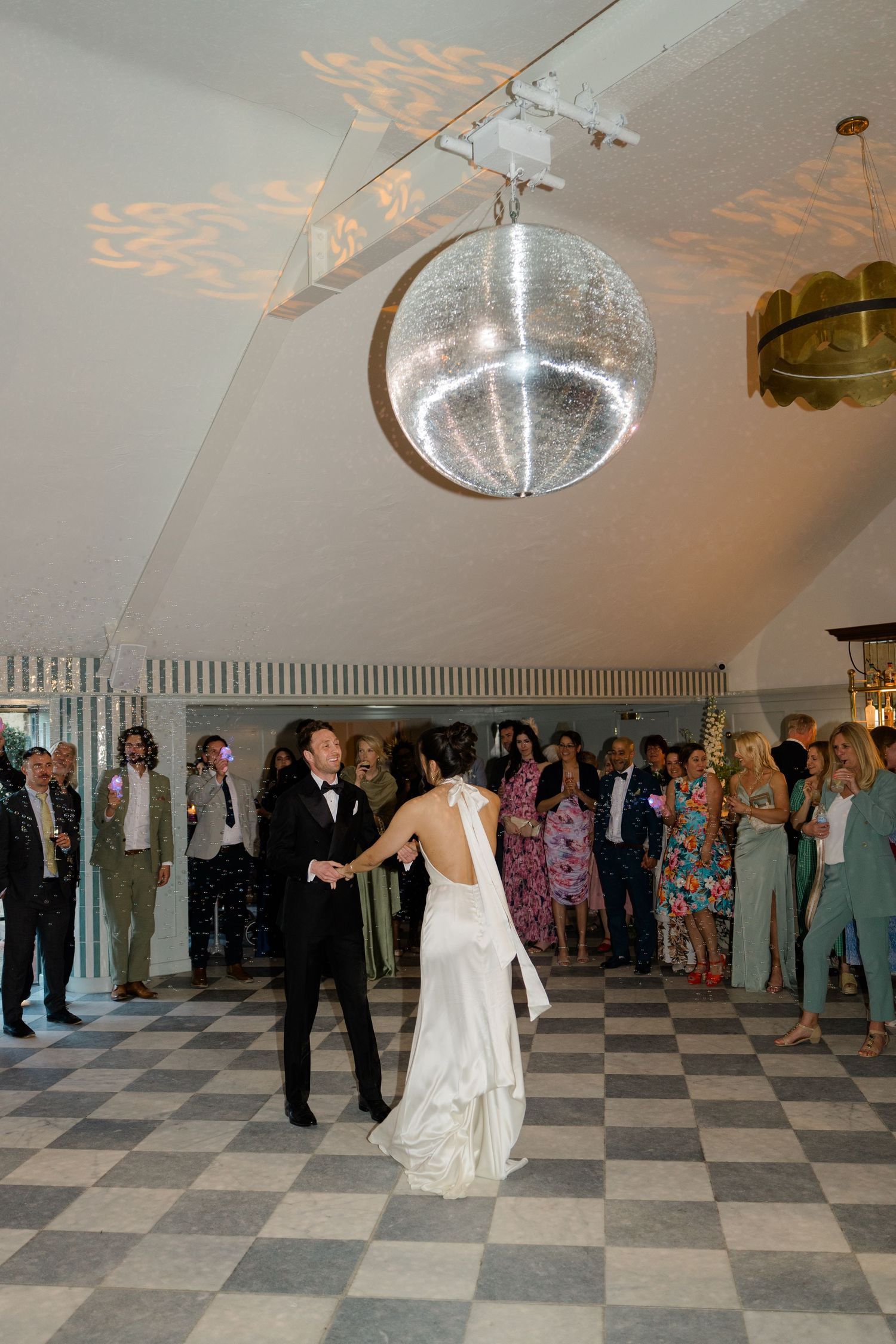 Bride and groom share first dance under a sparkling disco ball on checkered dance floor before guests.