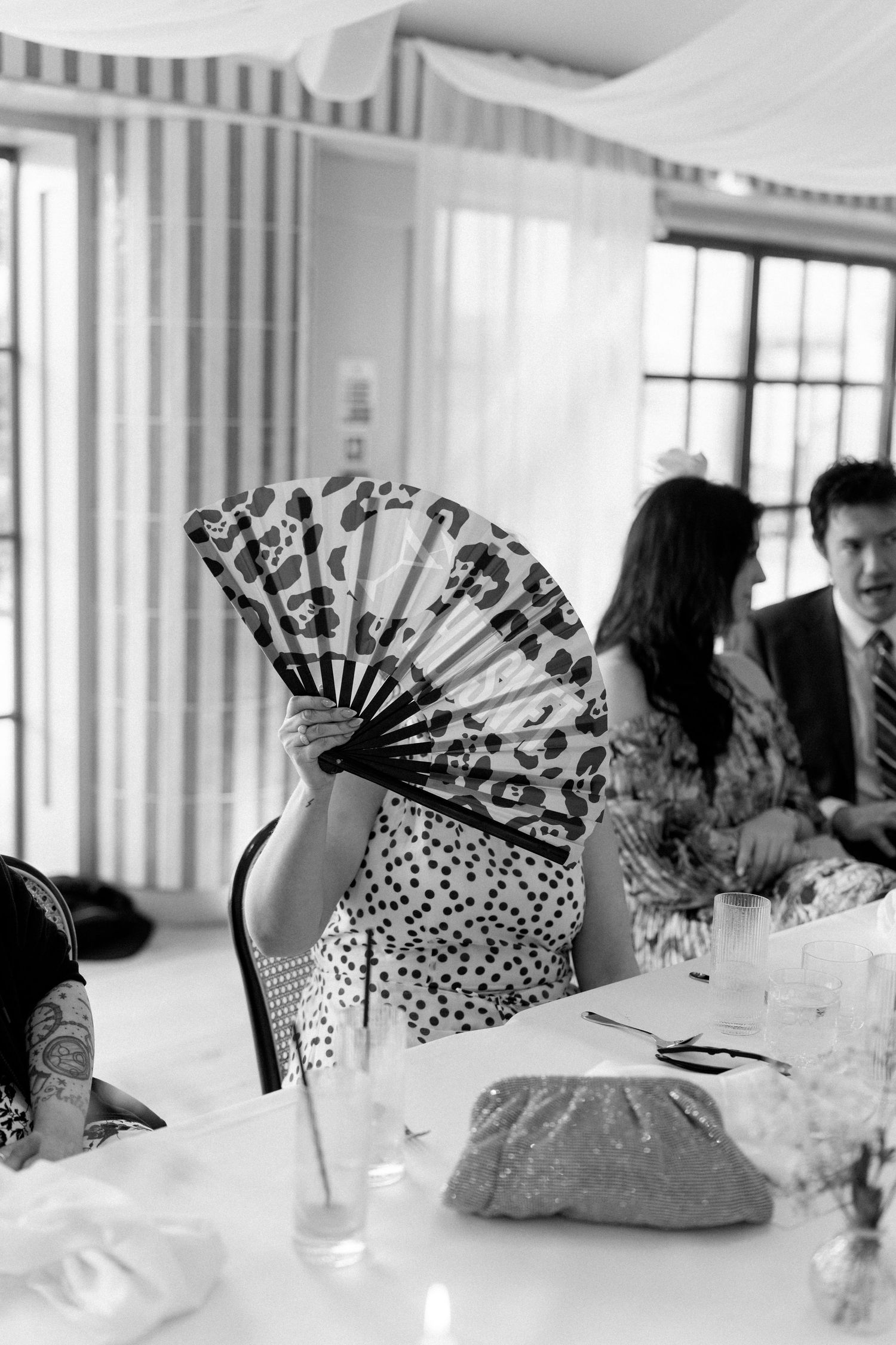 Black and white photo of wedding guest hiding behind leopard print fan at elegant reception table setting.