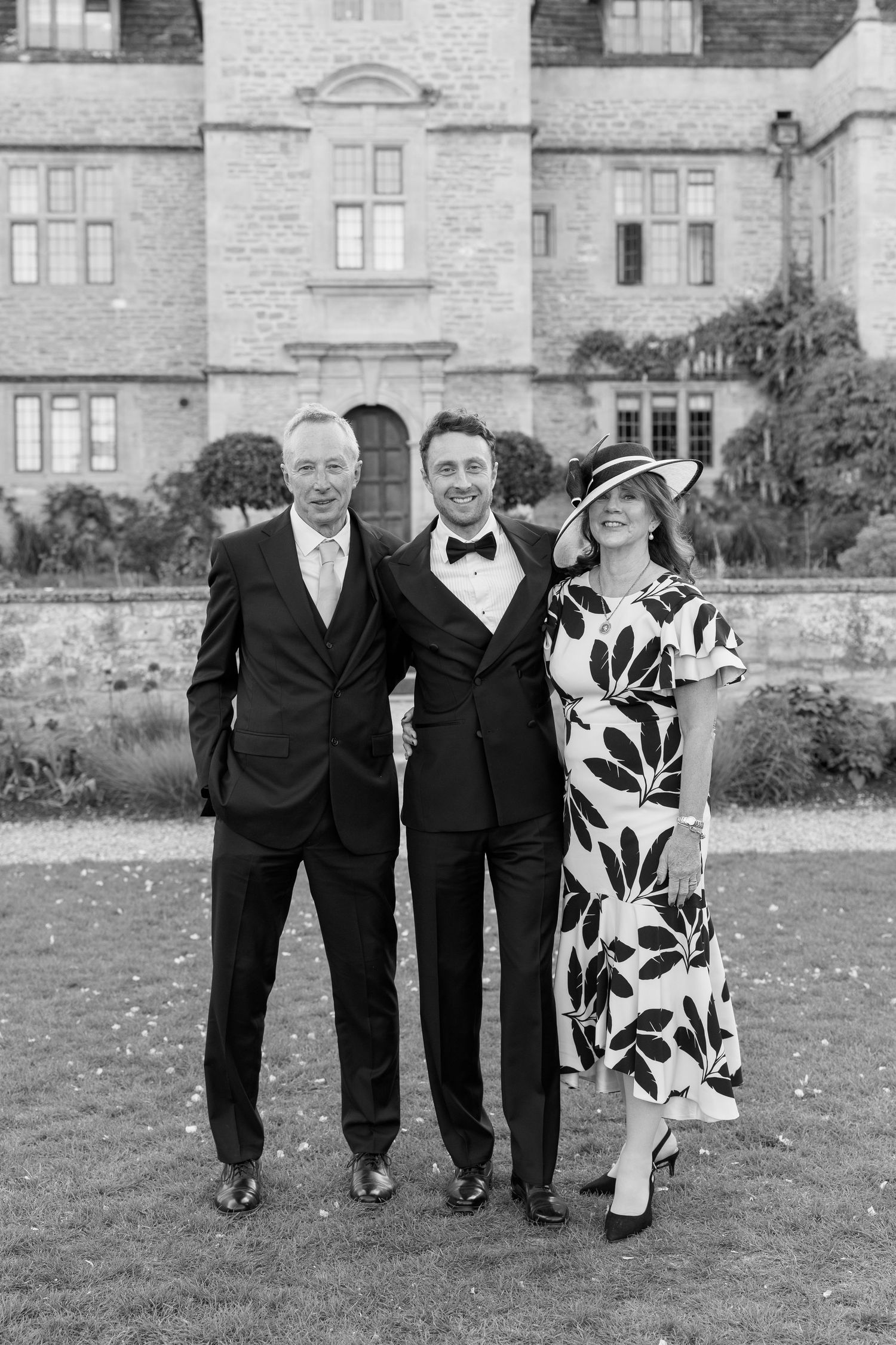 Groom poses with parents in black and white portrait outside a grand historic stone manor house.