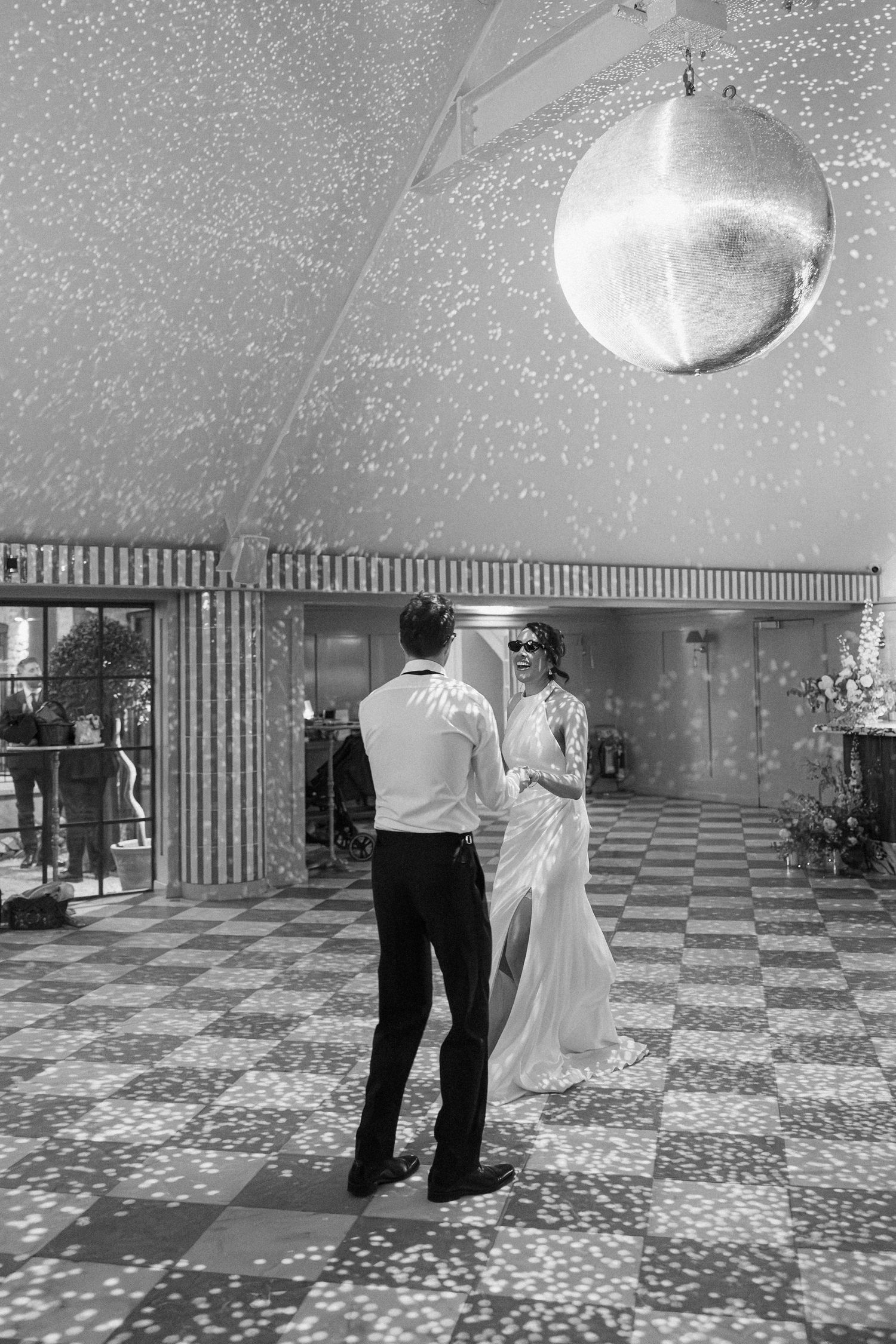 Black and white first dance photo under glittering disco ball on checkered dance floor at wedding.