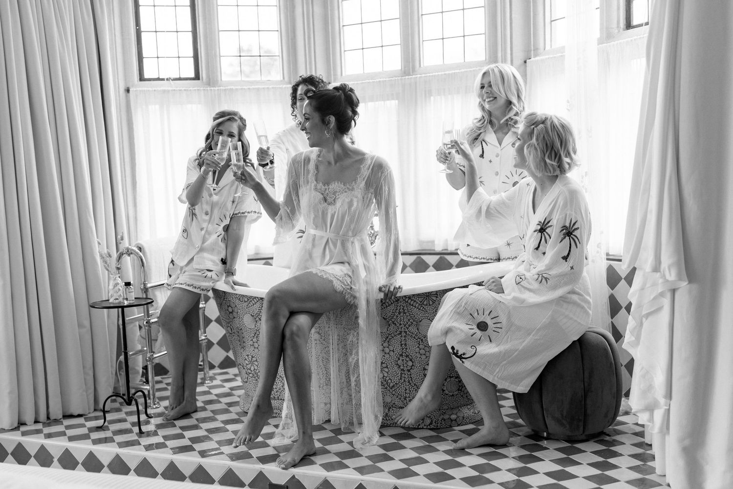 Black and white photo of bride and bridesmaids in robes laughing together by a window.