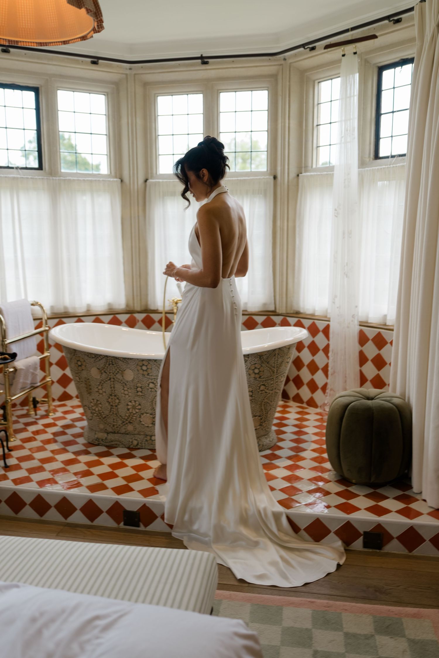 Bride in elegant backless white wedding dress stands by vintage bathtub in ornate tiled bathroom.