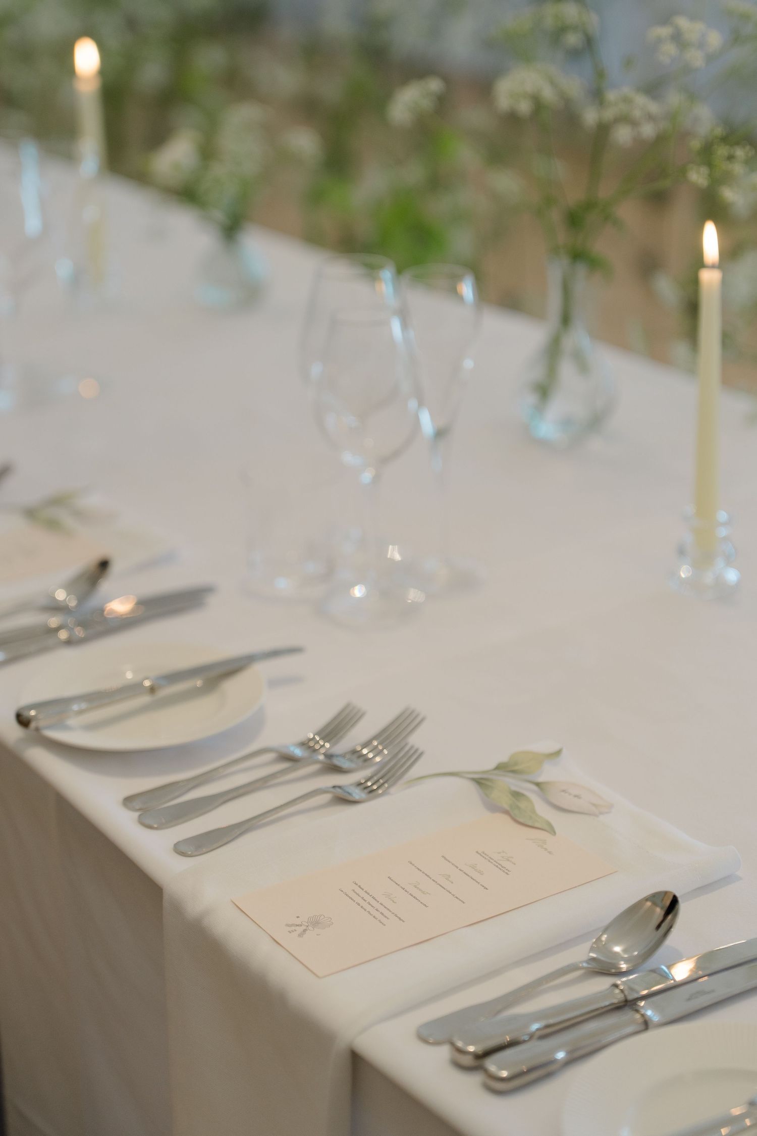 Close-up of elegant wedding table setting with silver cutlery, candles, glassware, and floral menu card.