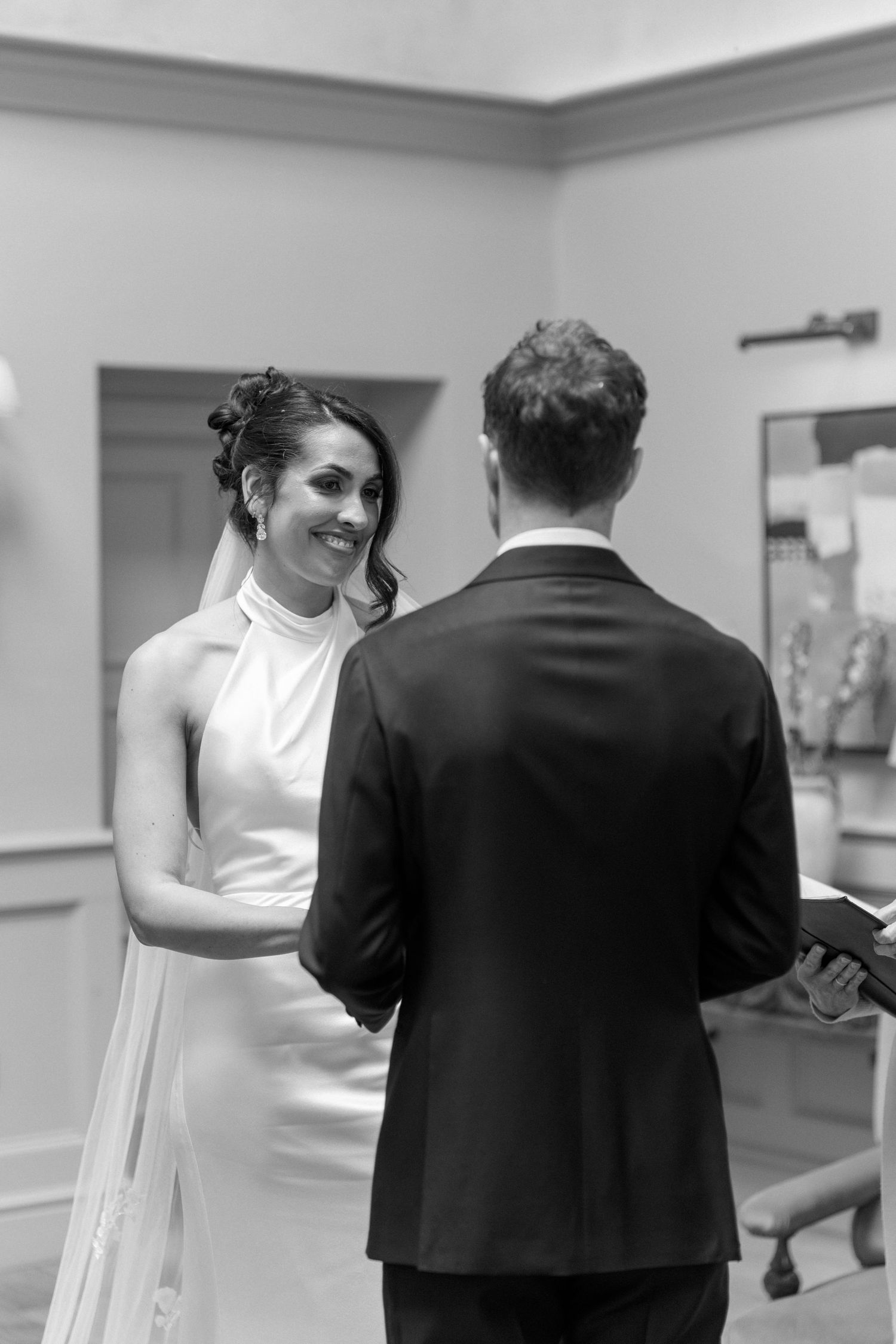 Smiling bride exchanges vows with groom during intimate indoor wedding ceremony in black and white.