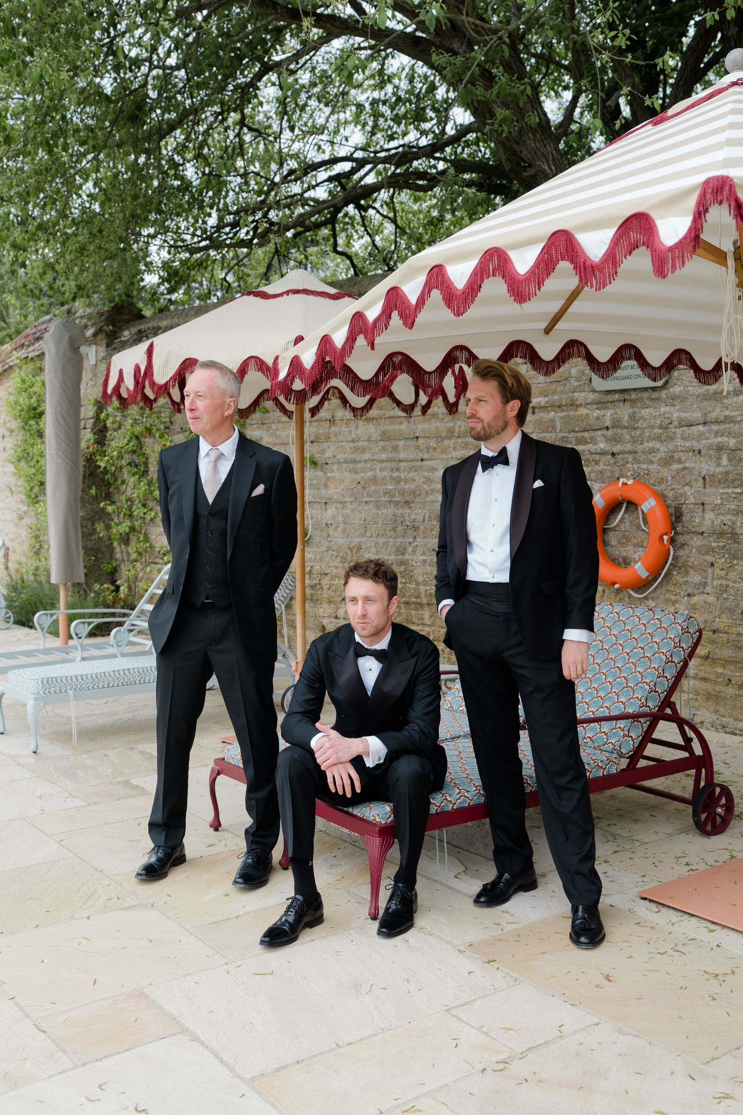Three groomsmen in black tuxedos posing under a striped canopy beside a swimming pool.